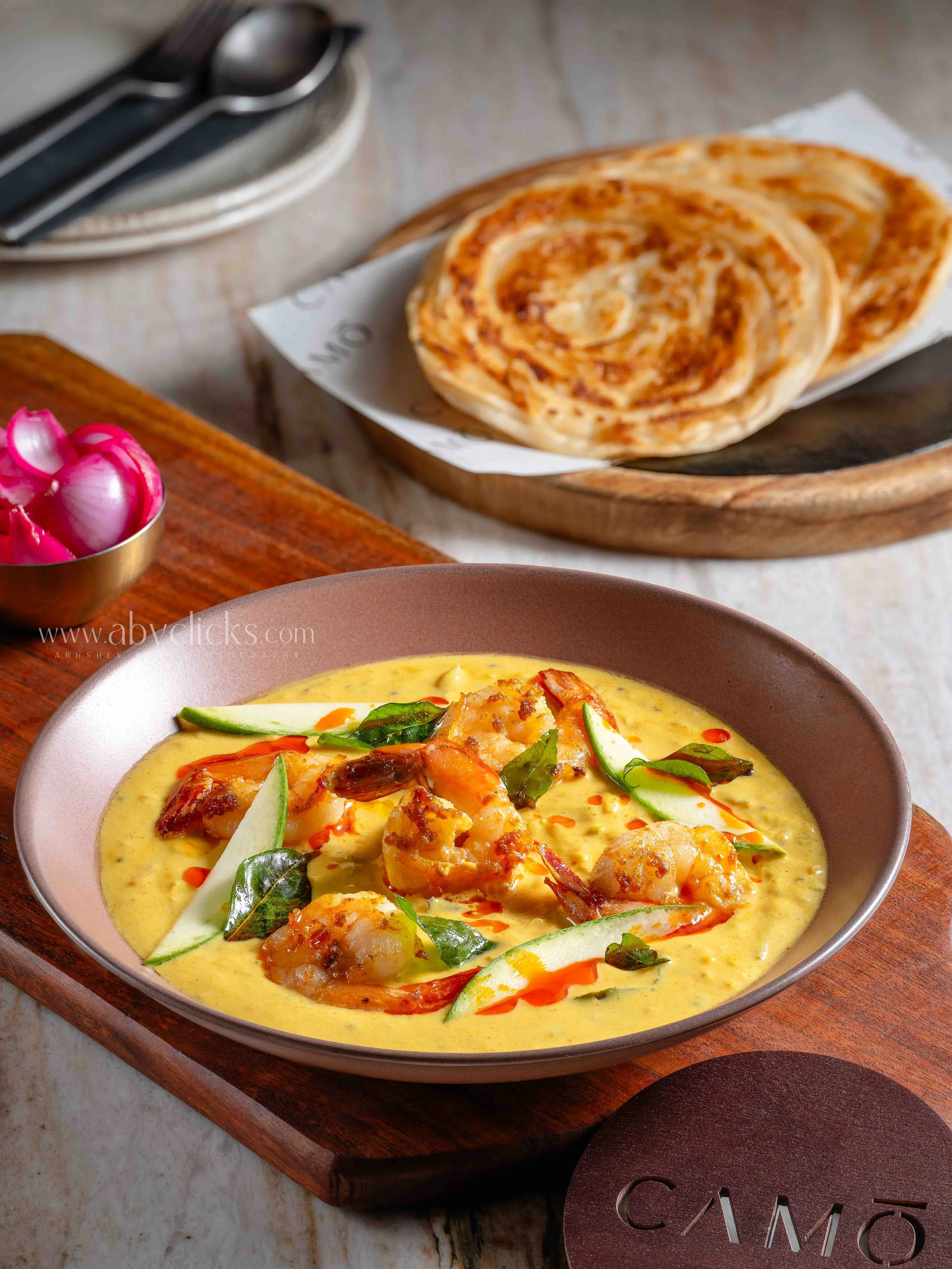 A bowl of yellow curry with shrimp, vegetables, and herbs, placed on a wooden serving board. In the background, naan bread on a paper-lined wooden tray, a small bowl of pickled onions, and some utensils on a plate.