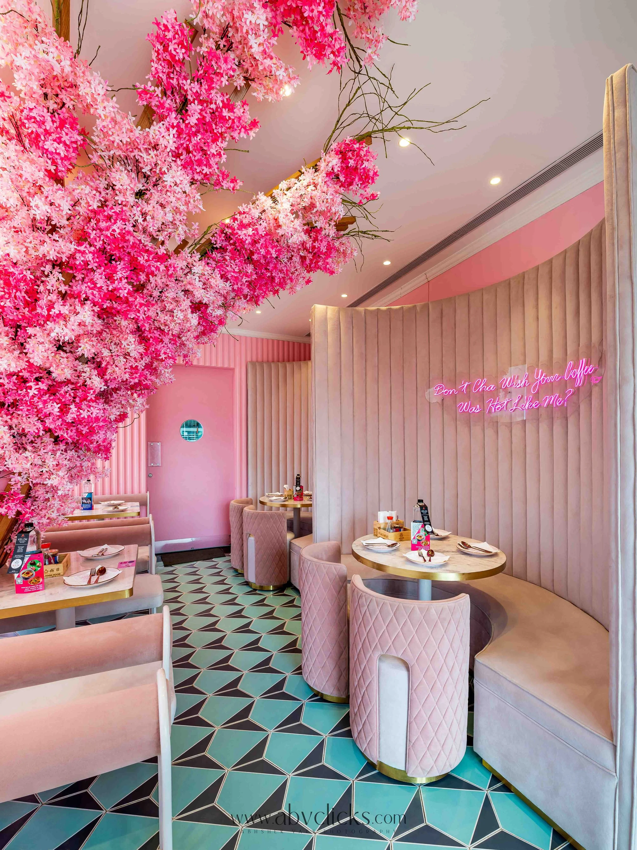 Pink and white floral decorations with pink upholstered seating, round tables set with dishes, on a patterned floor, in a pink themed interior with a neon sign reading 'Don't Cha Wish Your Coffee Was Hot Like Me?'