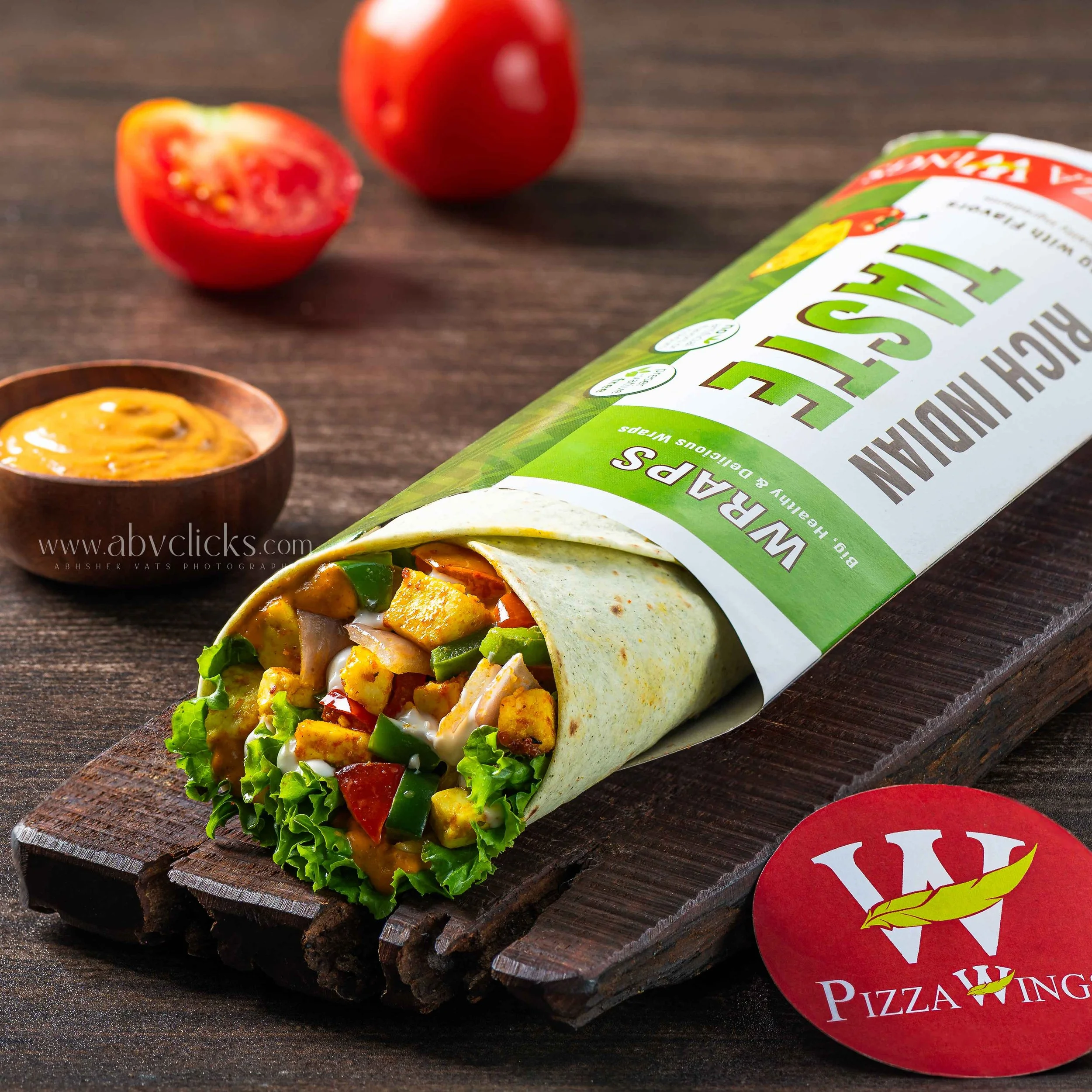 A veggie wrap with lettuce, peppers, and chicken on a wooden board, with fresh tomato slices and a pizza box in the background.