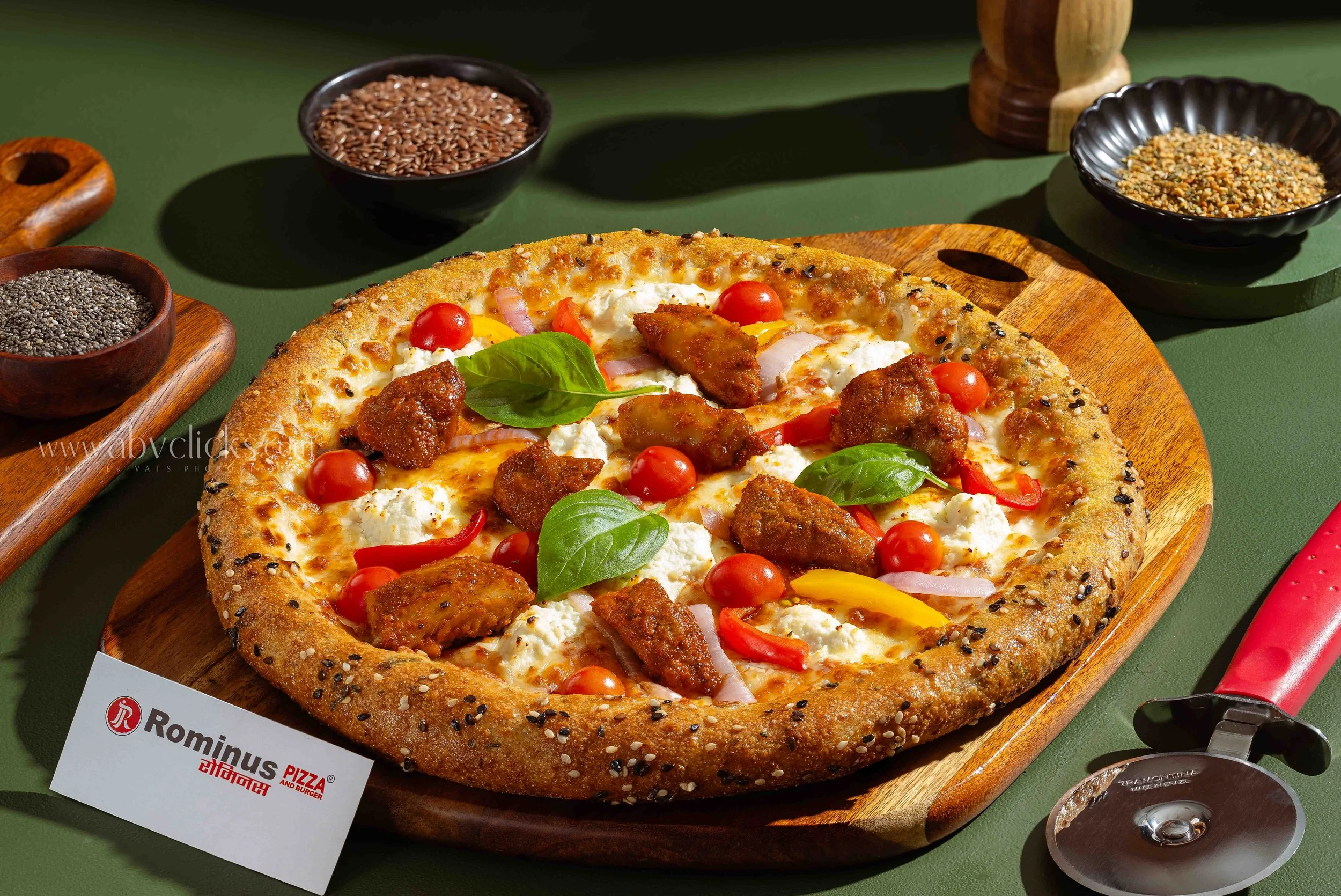 A pizza with a thick crust topped with cherry tomatoes, herbs, cheese, onions, and sliced meat, placed on a wooden serving board with containers of various seeds around it.