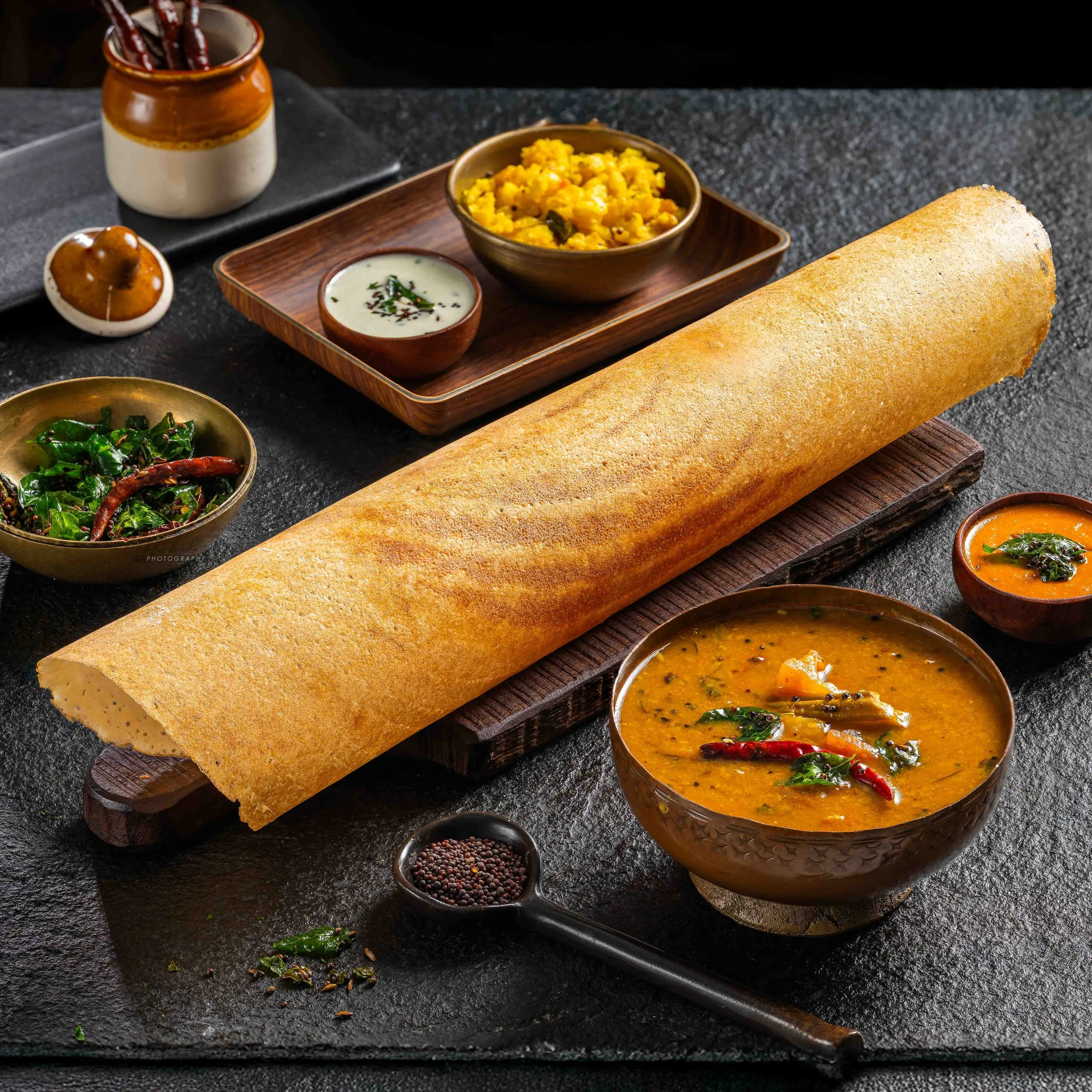 Assorted South Indian dishe on a dark surface, including a large dosa, rice dish, sambhar, chutney, and spices.