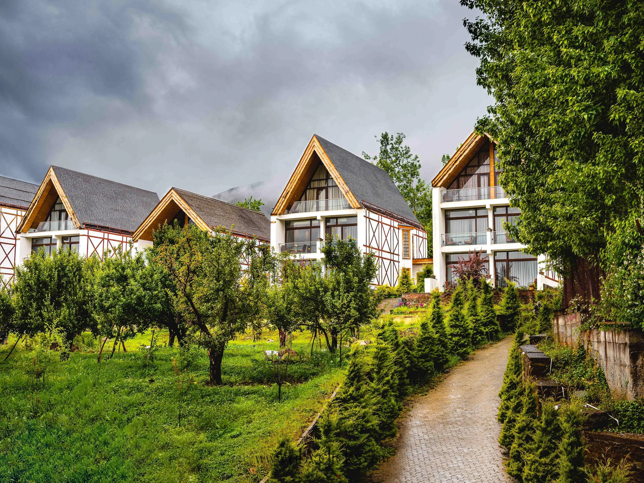 View of chalet-style buildings with steep roofs, surrounded by lush greenery and trees, under a cloudy sky.