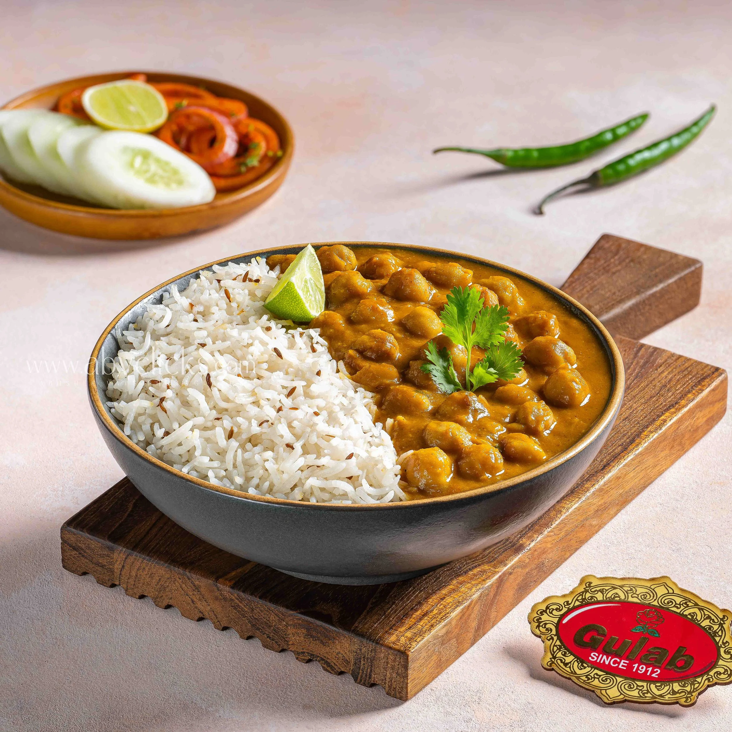 A bowl of white rice and chickpea curry garnished with cilantro and lime, with a side dish of pickled vegetables and lemon wedge in a wooden bowl, on a wooden tray with a decorative paper beneath.