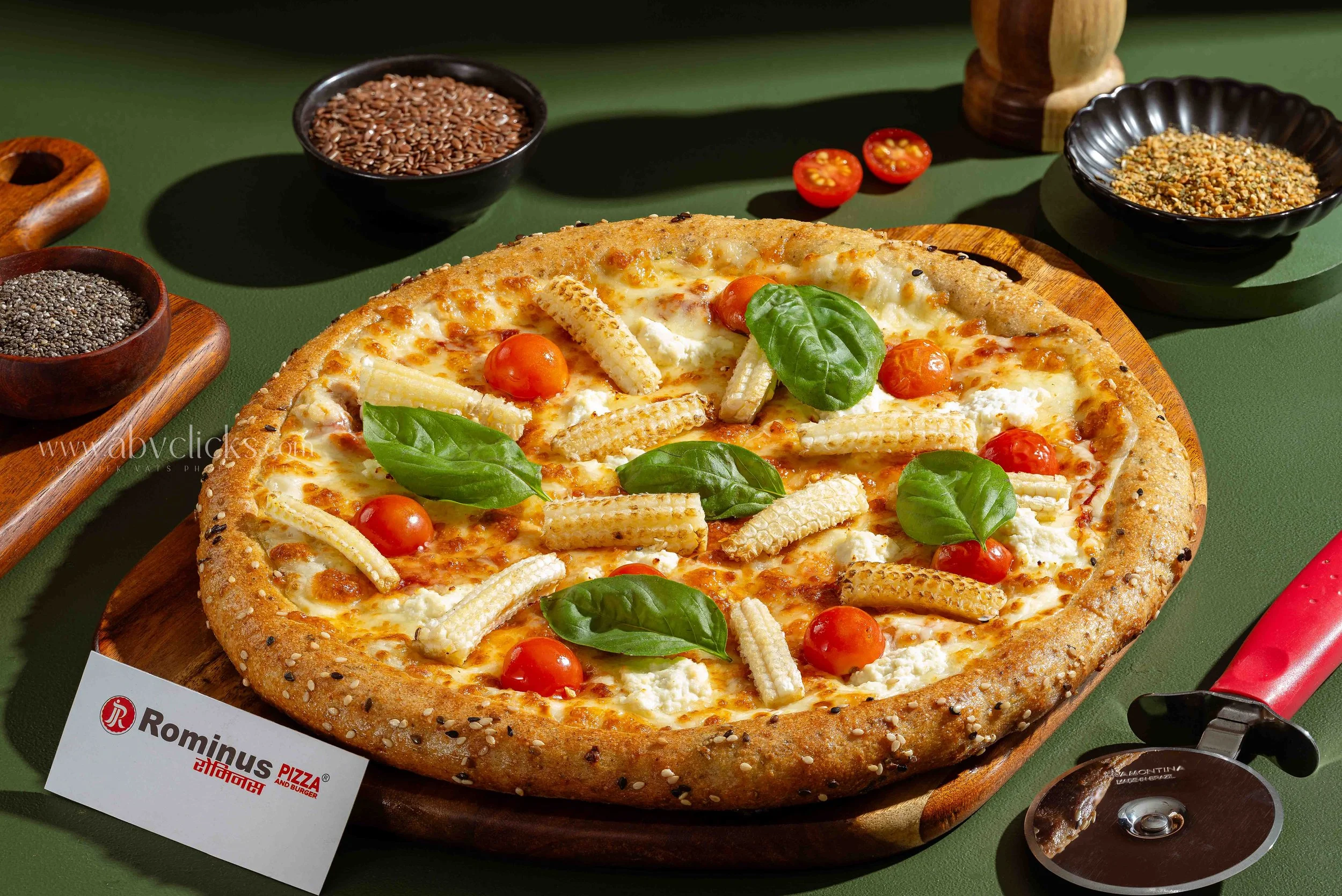 A pizza with cherry tomatoes, basil leaves, and cheese on a wooden board surrounded by small bowls of seeds and crushed nuts, with a pizza cutter nearby.