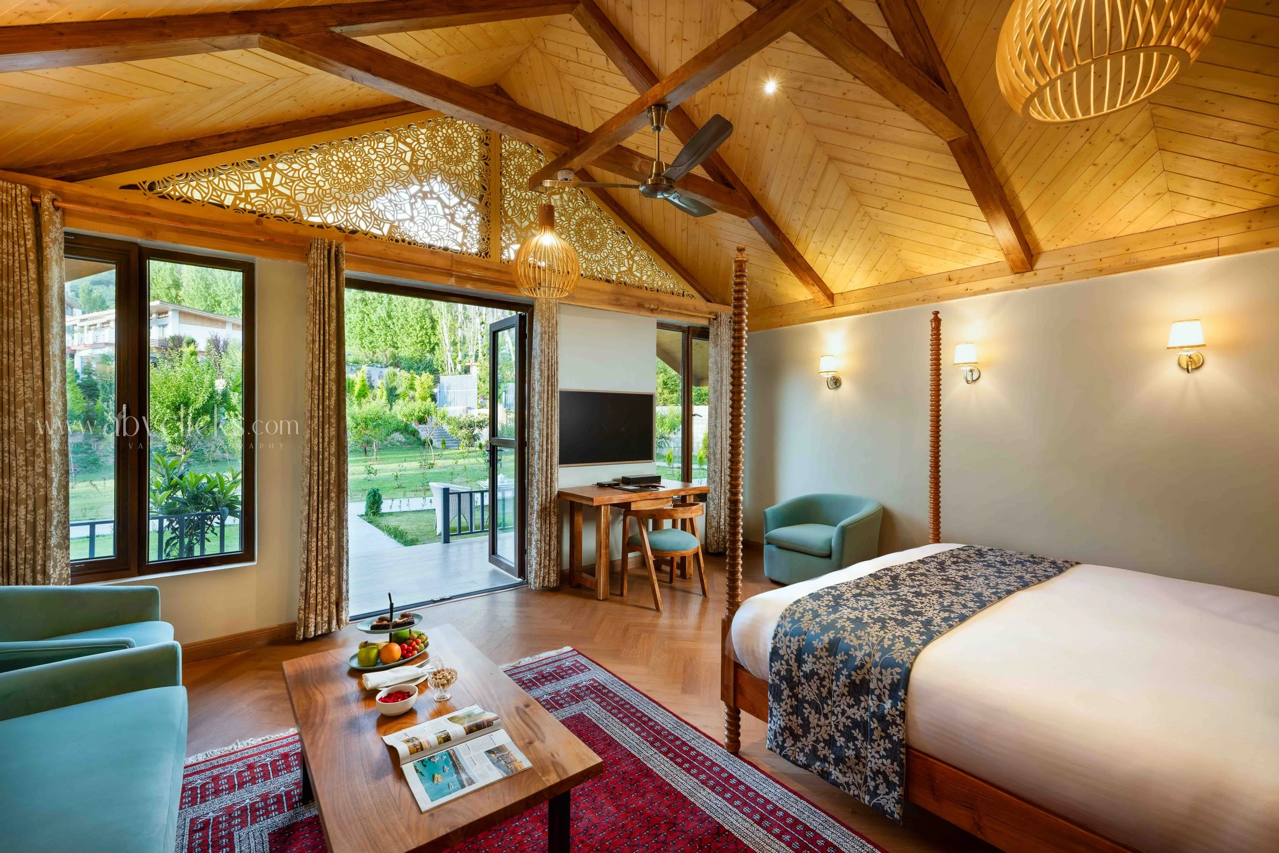 A cozy bedroom with a wooden ceiling and large windows opening to a garden view, featuring a bed, armchair, small dining table, and various lighting fixtures.