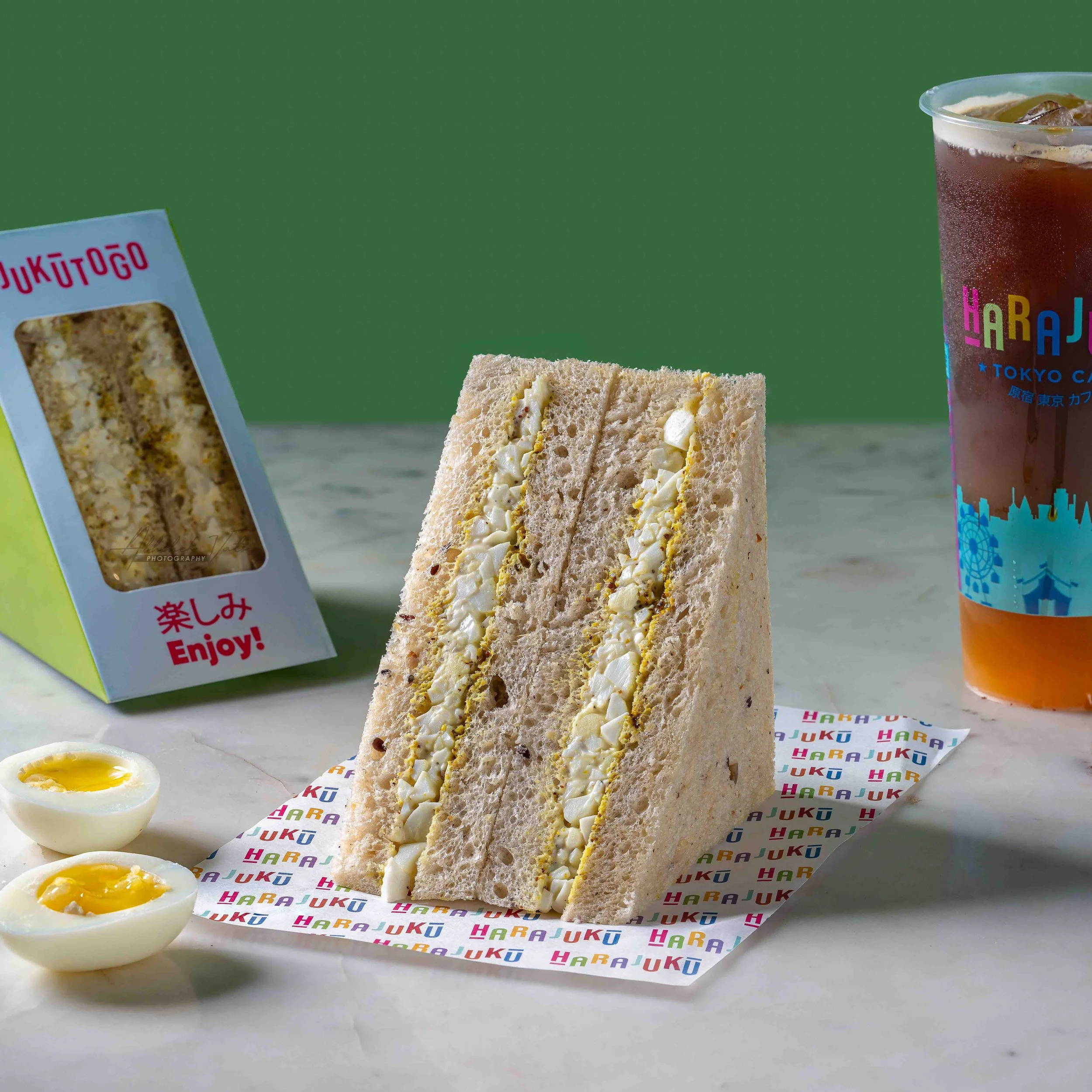 Close-up of a sandwich with light bread and mayonnaise filling, sliced in half, with a soft-boiled egg in a cup on the side, on a surface with colorful alphabet paper and a beverage in a clear plastic cup with ice, against a green background.
