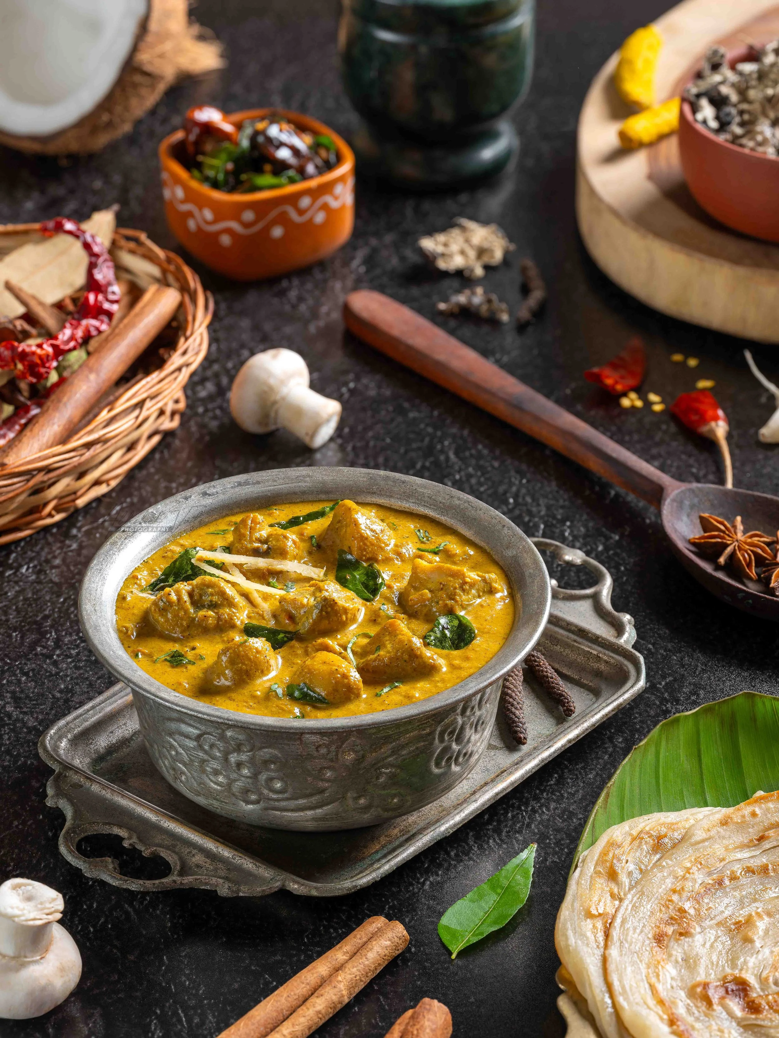 A bowl of Indian chicken curry garnished with herbs, surrounded by spices, herbs, and ingredients on a black surface.