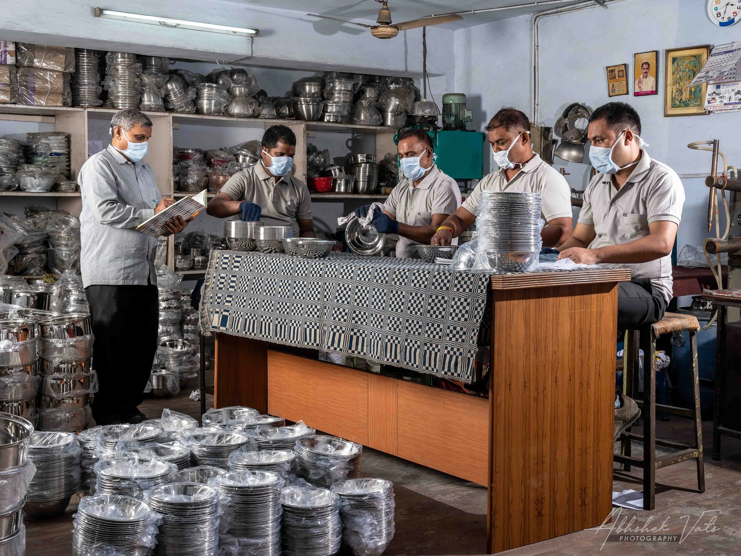 A group of five men in masks and gloves working with stainless steel kitchenware in a shop. One man is reading a book, while the others are arranging and inspecting the metal utensils on the table. Shelves filled with more kitchenware are visible in 