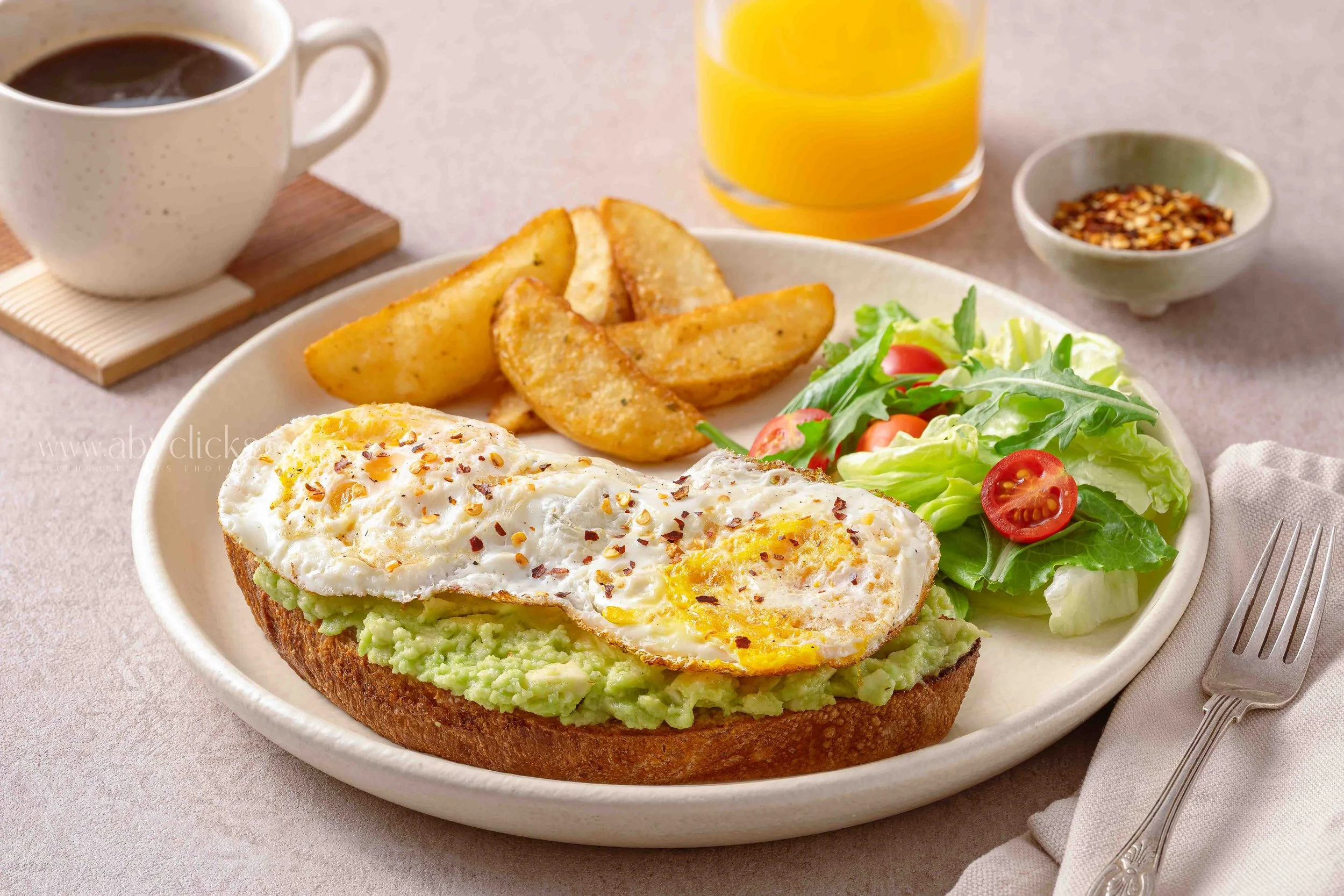 A breakfast plate with avocado toast topped with fried eggs, potato wedges, a side salad with cherry tomatoes and greens, a glass of orange juice, a cup of black coffee, and a small dish of red pepper flakes.