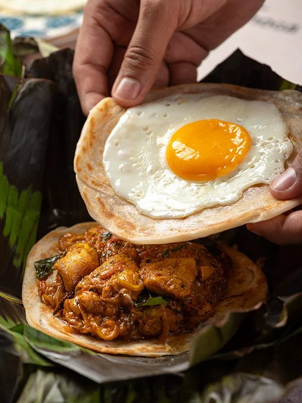Hand placing a fried egg on top of a bowl of spicy chicken curry in a banana leaf basket.
