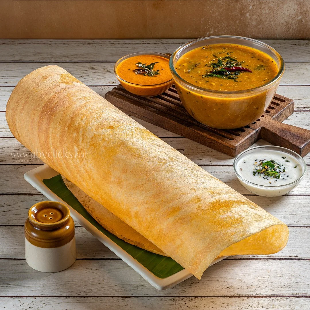 A large dosa (Indian pancake) served on a white rectangular plate with a green leaf underneath. Three bowls of different Indian curries or chutneys are nearby, including sambar and coconut chutney, garnished with herbs and spices. The dishes are on a