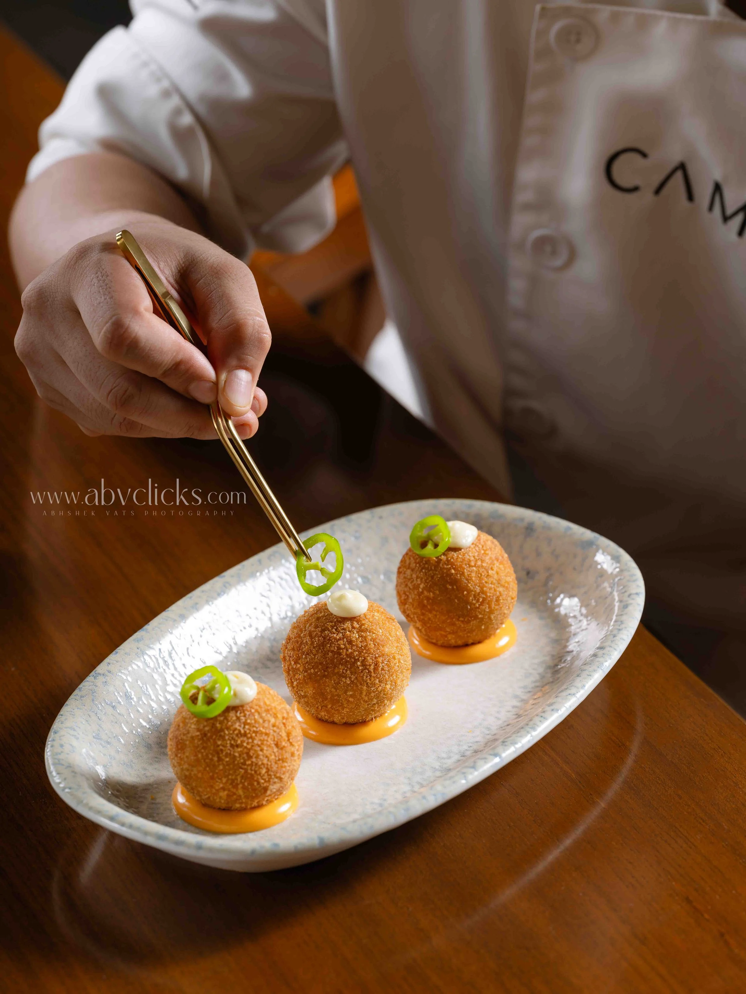 Three golden-brown, round, fried appetizers on a white oval plate with a speckled pattern, garnished with small slices of green pepper and dollops of white sauce on top, served on a wooden table.