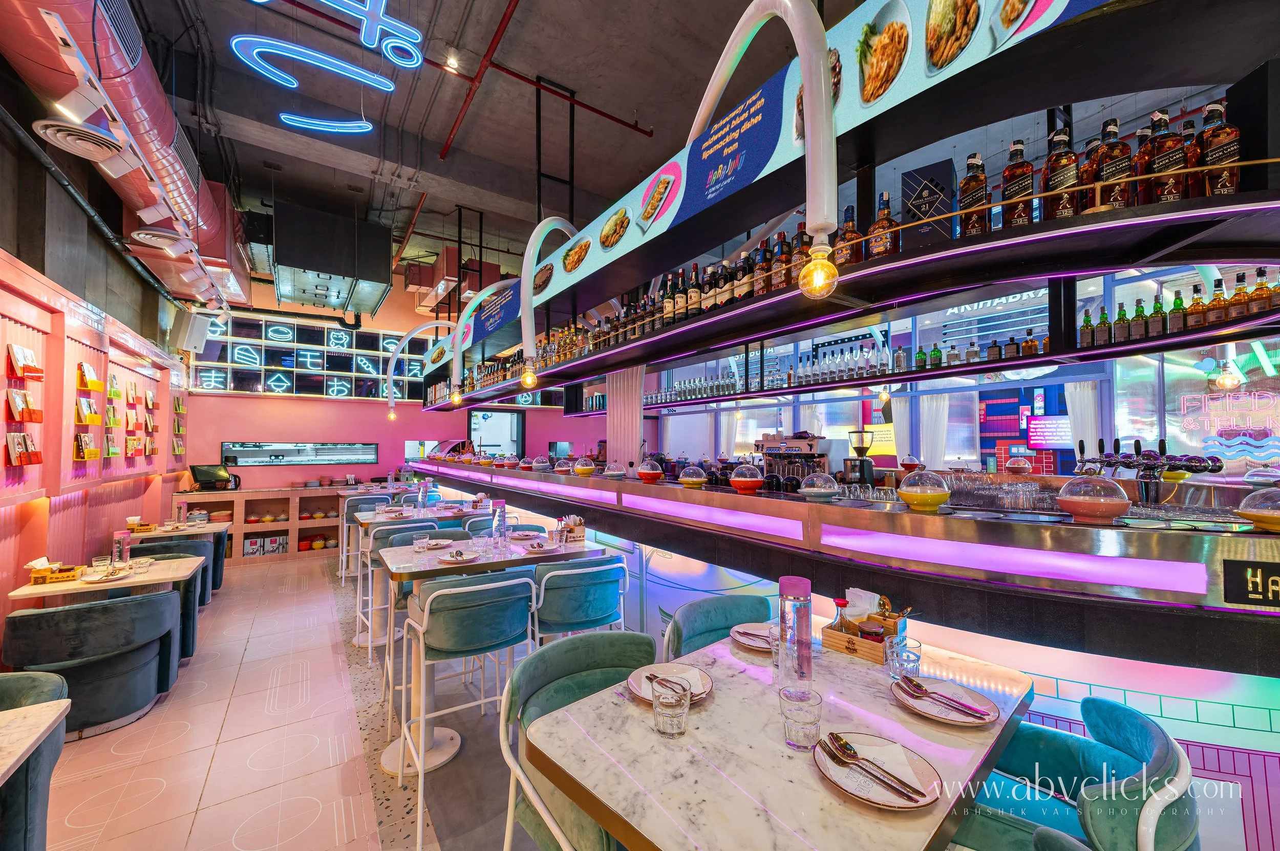 Interior of a vibrant, modern restaurant with pink walls, neon lights, and a long bar counter accentuated with purple lighting. There are tables set with glassware and plates, and shelves with colorful books and bottles.