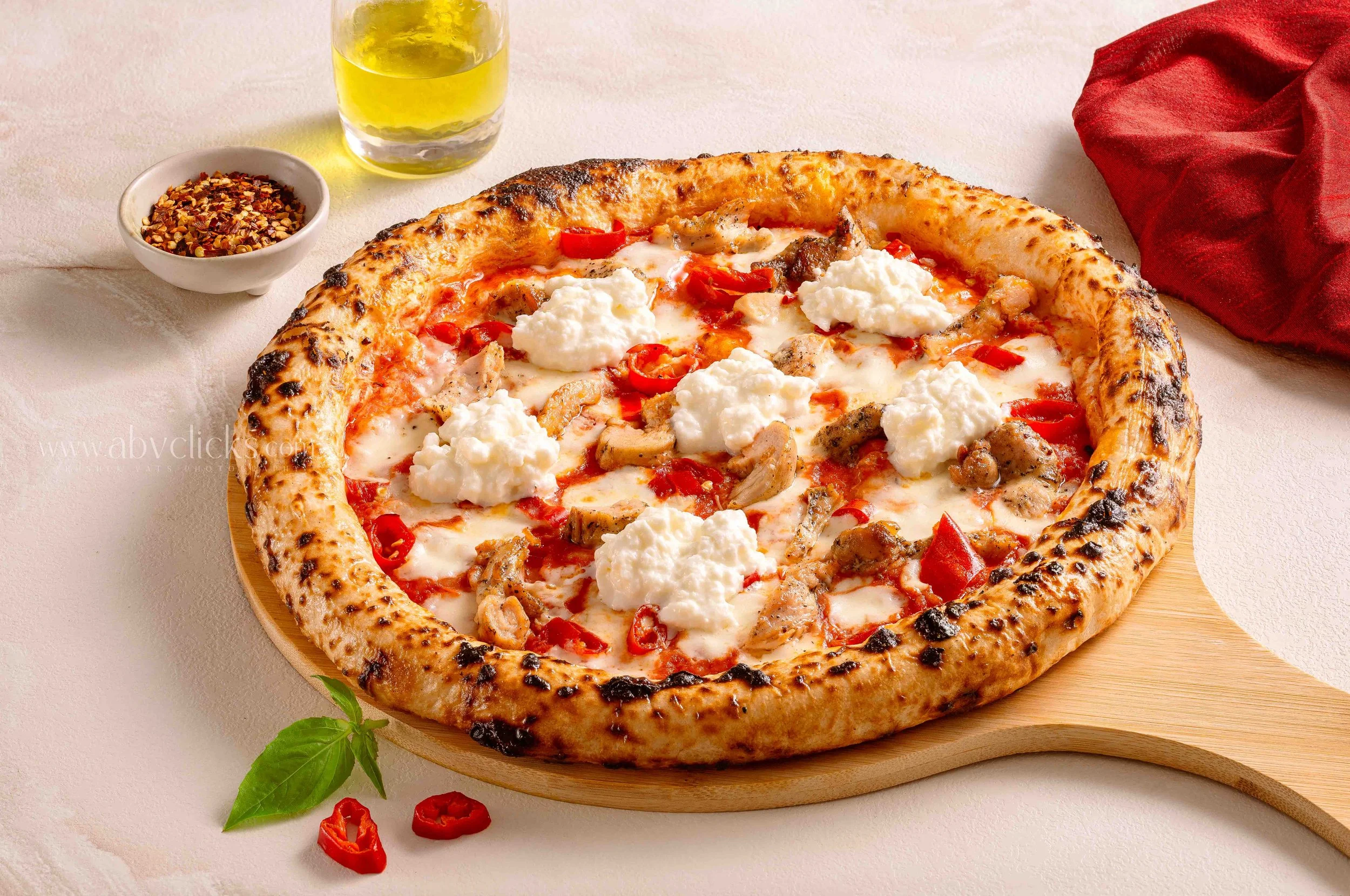 A pizza with a thick crust topped with cheese, red peppers, and chicken on a wooden board, with a glass of yellow beverage, a bowl of crushed red pepper flakes, and a red napkin nearby.