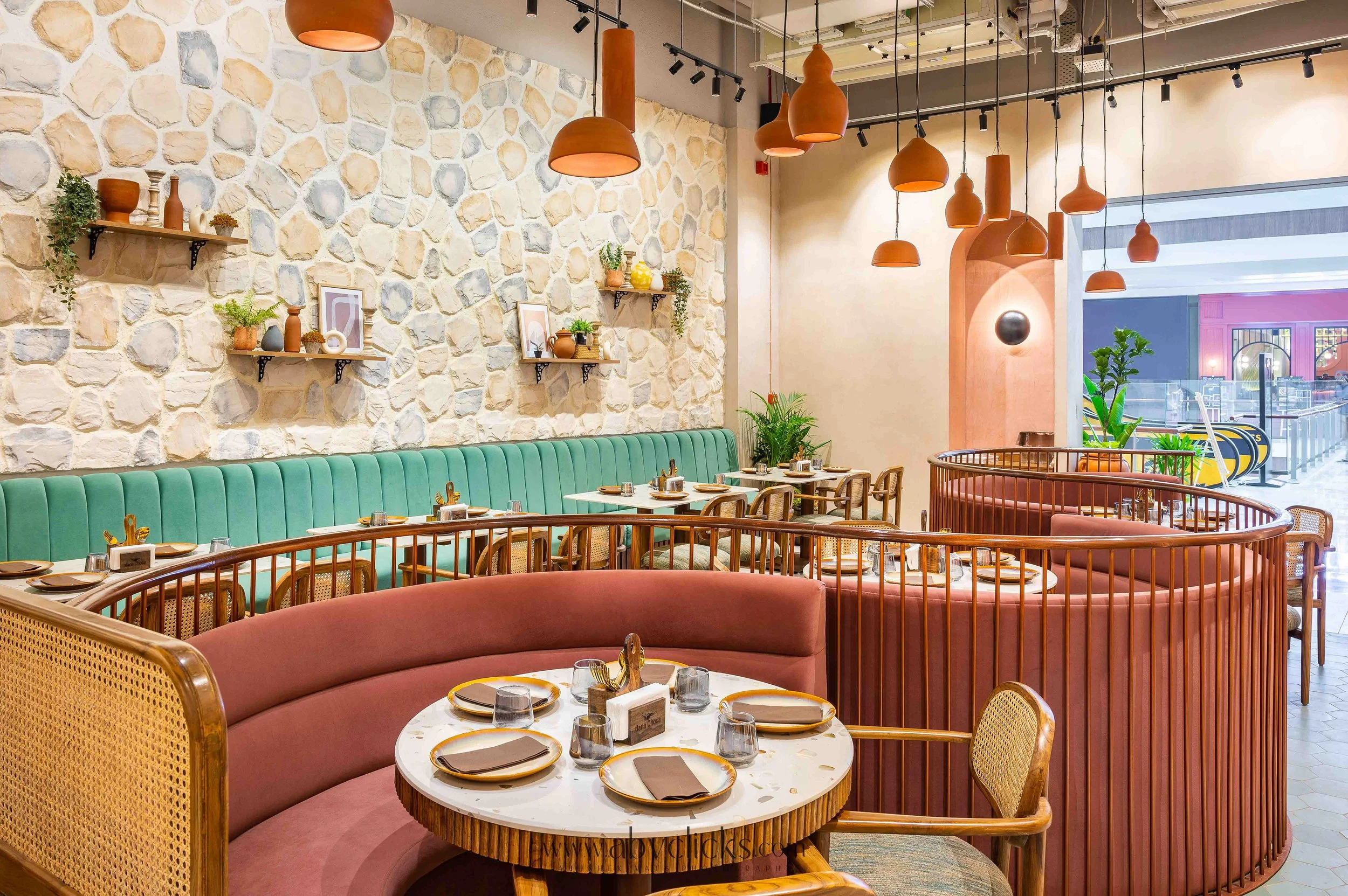 Interior of a restaurant with a stone accent wall, wooden shelves with vases and plants, hanging orange pendant lights, and a mix of green and pink padded booths with tables set for dining.