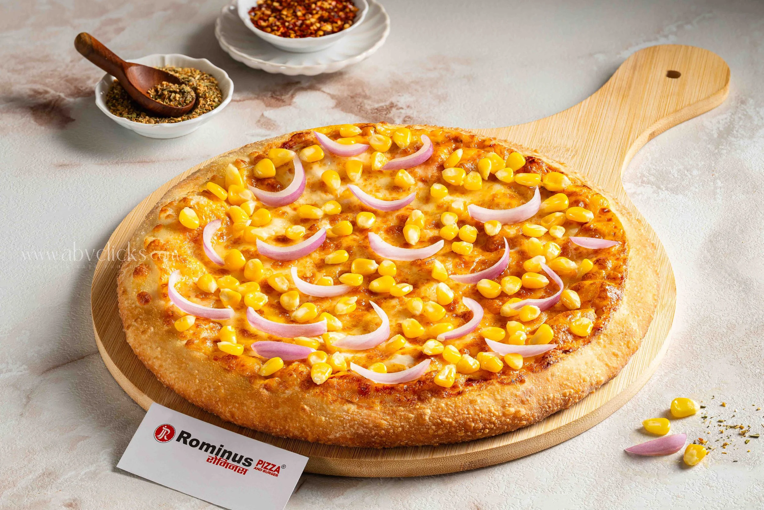 A pizza with cheese, corn, and onion slices on a wooden serving board, with small bowls of chili flakes and herbs in the background.