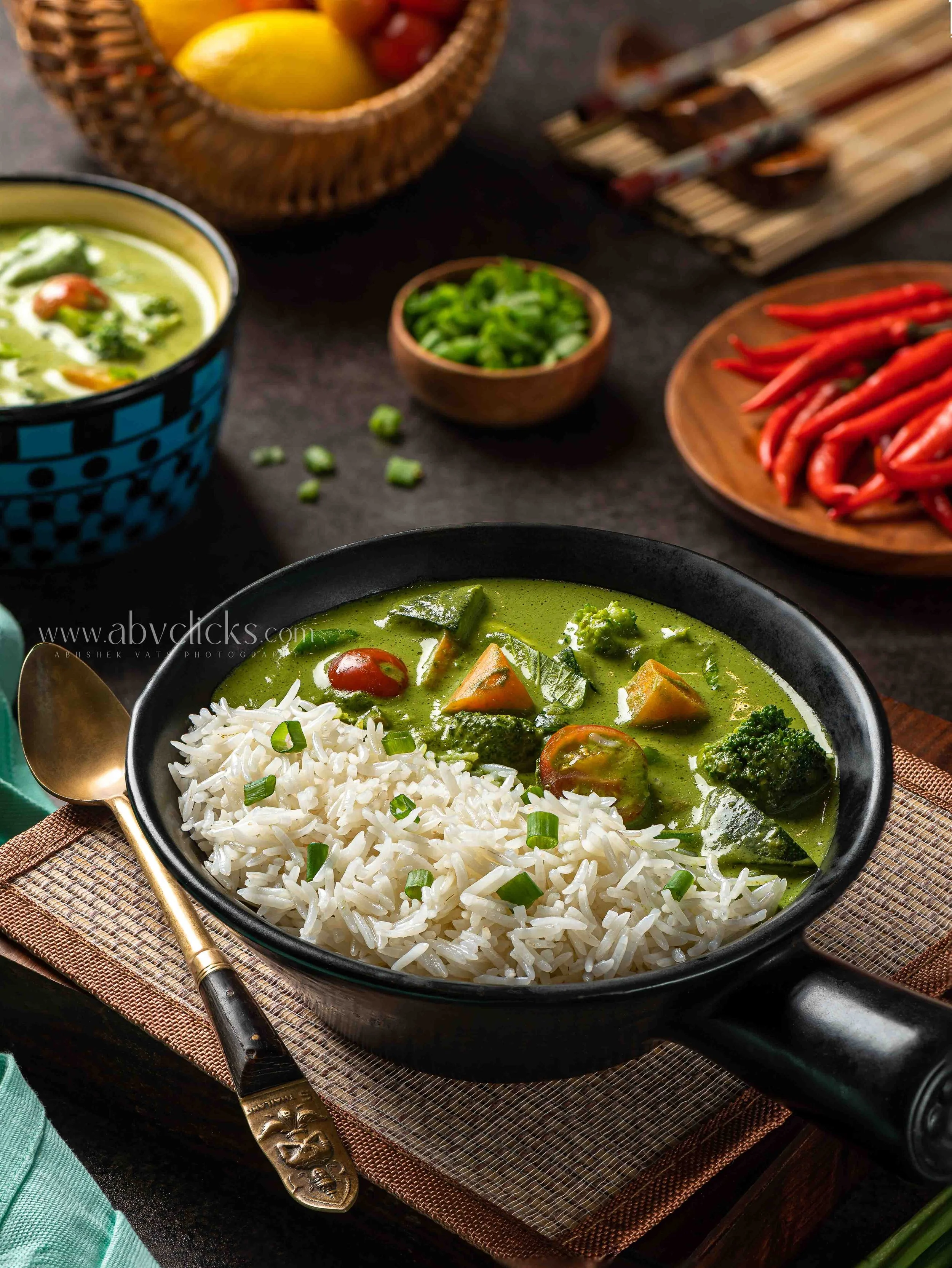 A bowl of green curry with vegetables and rice, accompanied by small bowls of chopped green onions and red chili peppers, on a dark table with a textured placemat and a gold spoon.