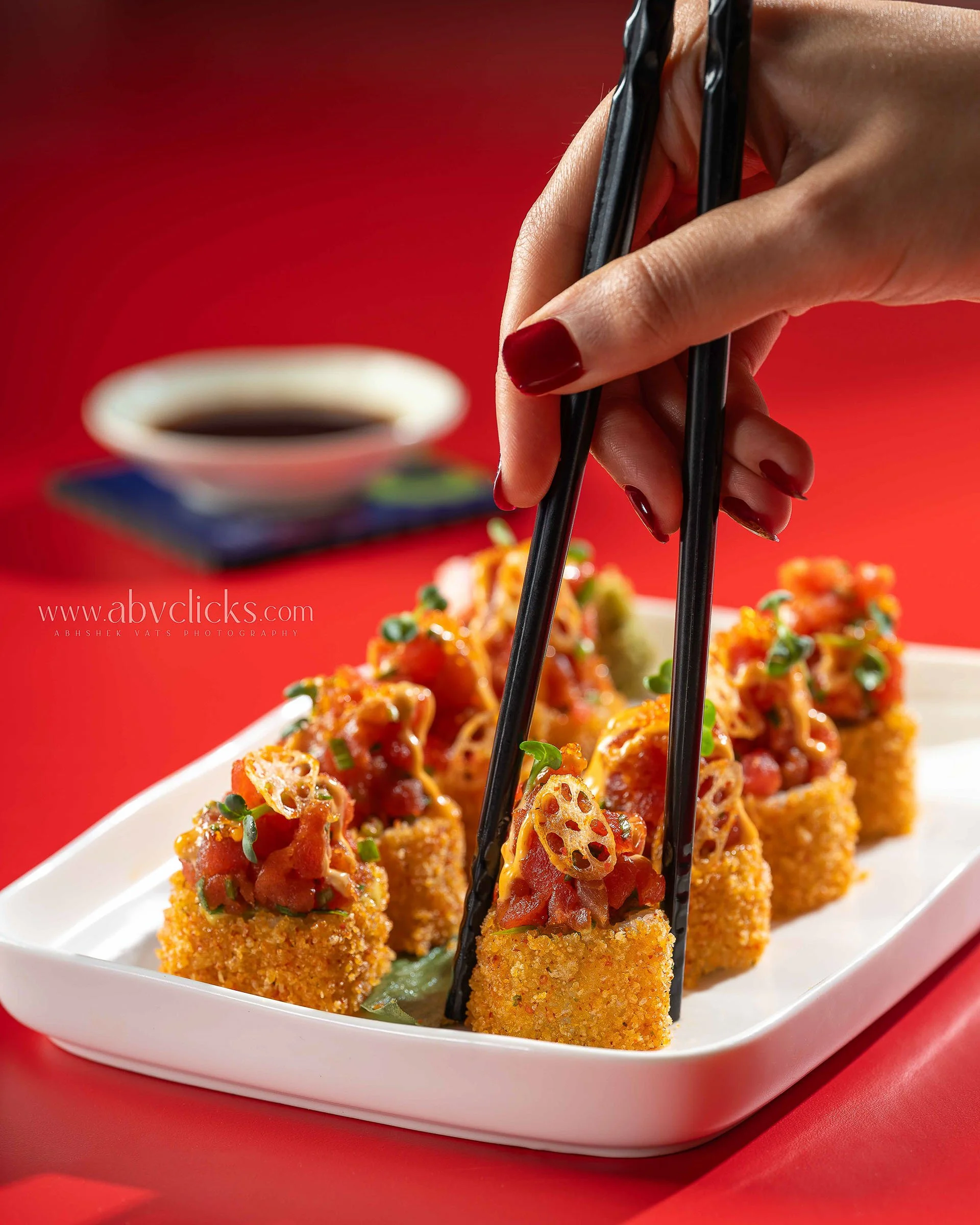 Close-up of crispy breaded appetizers topped with diced tomatoes, chopped green onions, and a sauce, being picked up with black chopsticks on a white dish with a red background.