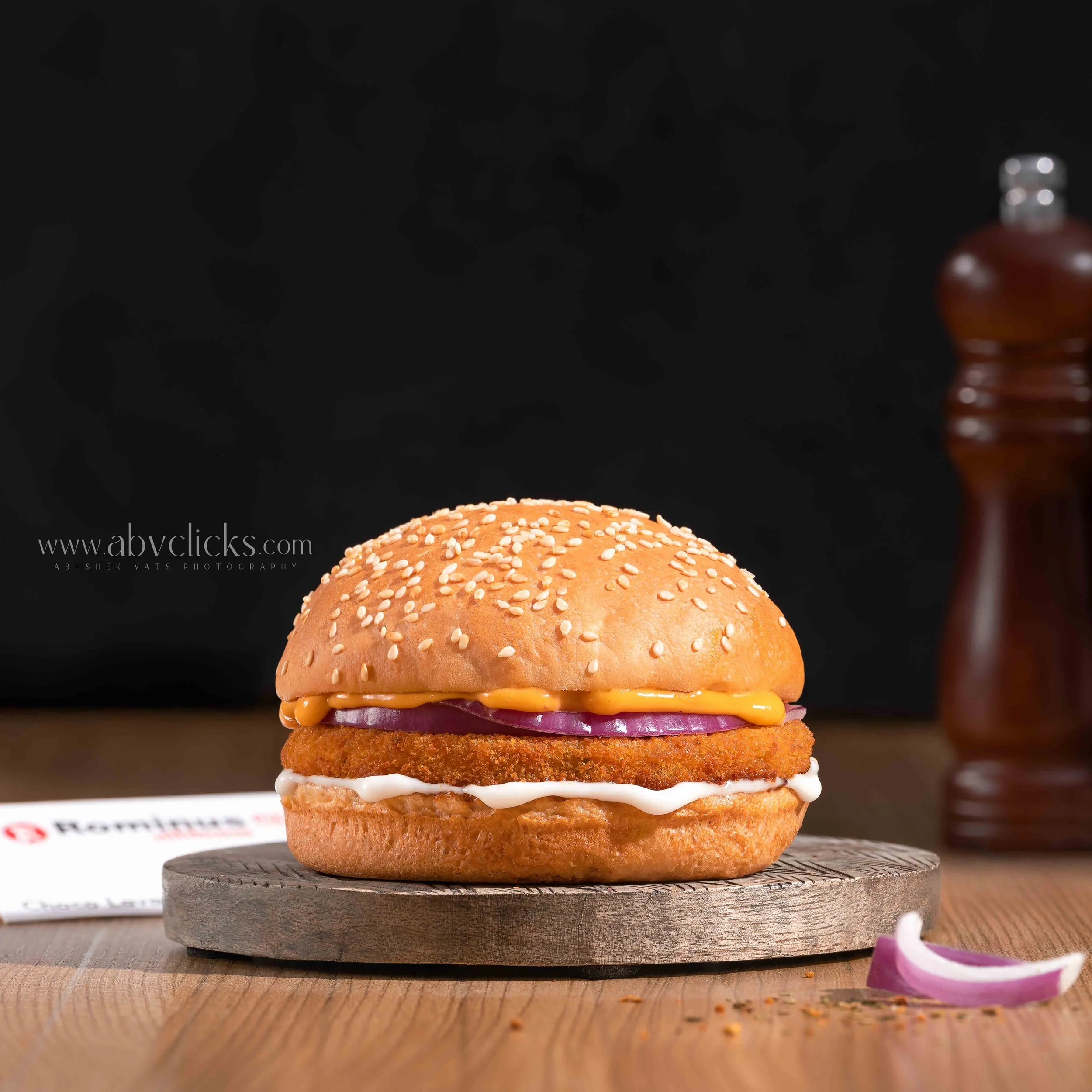 Close-up of a sesame seed burger bun on a wooden surface, with onion rings and a bottle of sauce in the background.