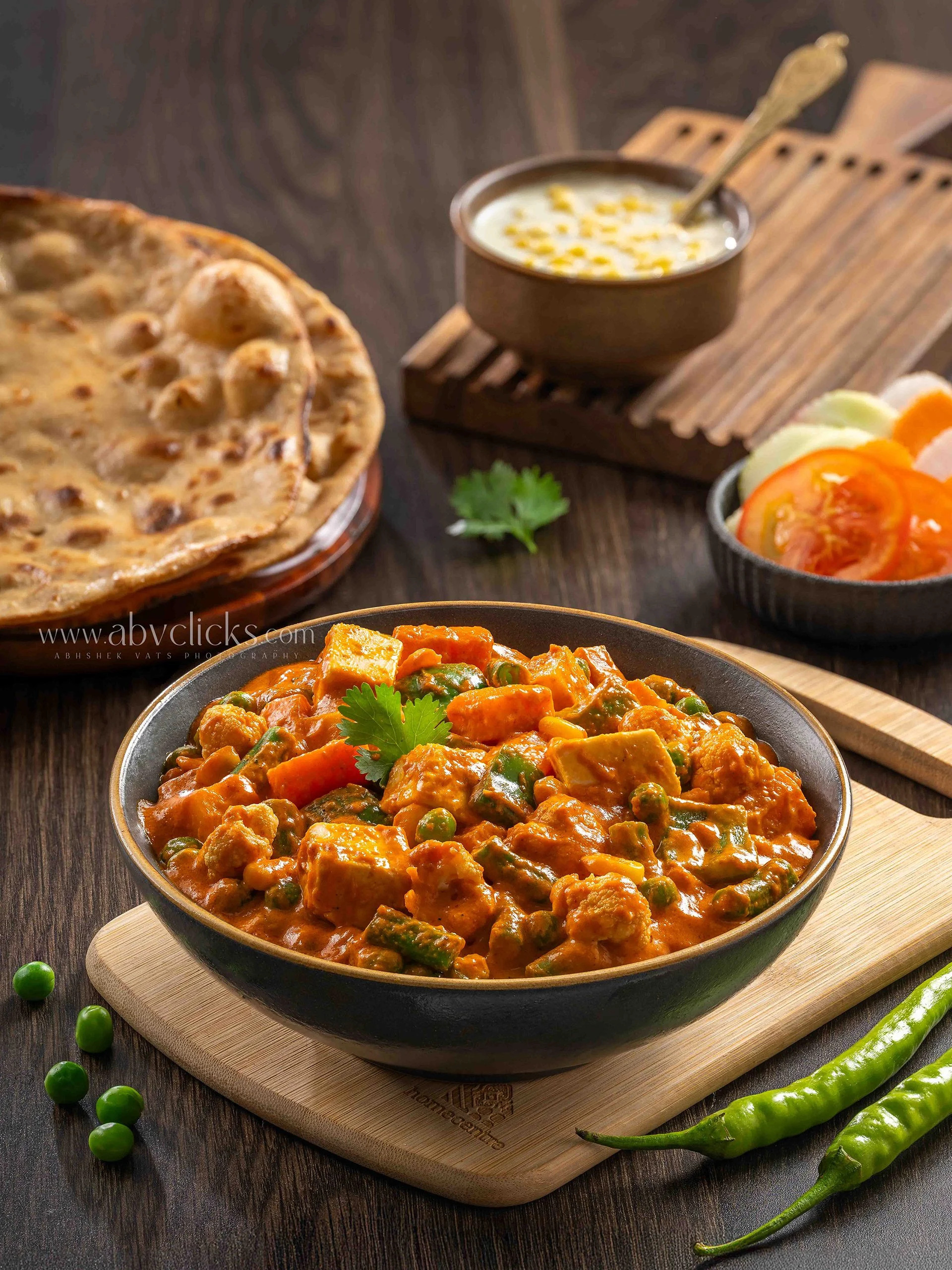 A bowl of paneer curry garnished with cilantro, placed on a wooden board with some green chilies and green peas around it, and a bowl of vegetables and salad in the background.