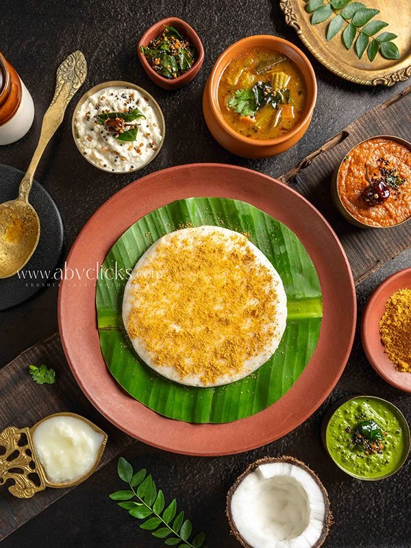 Variety of Indian dishes on a dark table, including a large plate with a round, layered snack garnished with a green leaf, accompanied by small bowls of different curries, chutneys, and sauces, along with a coconut and some leafy plants.