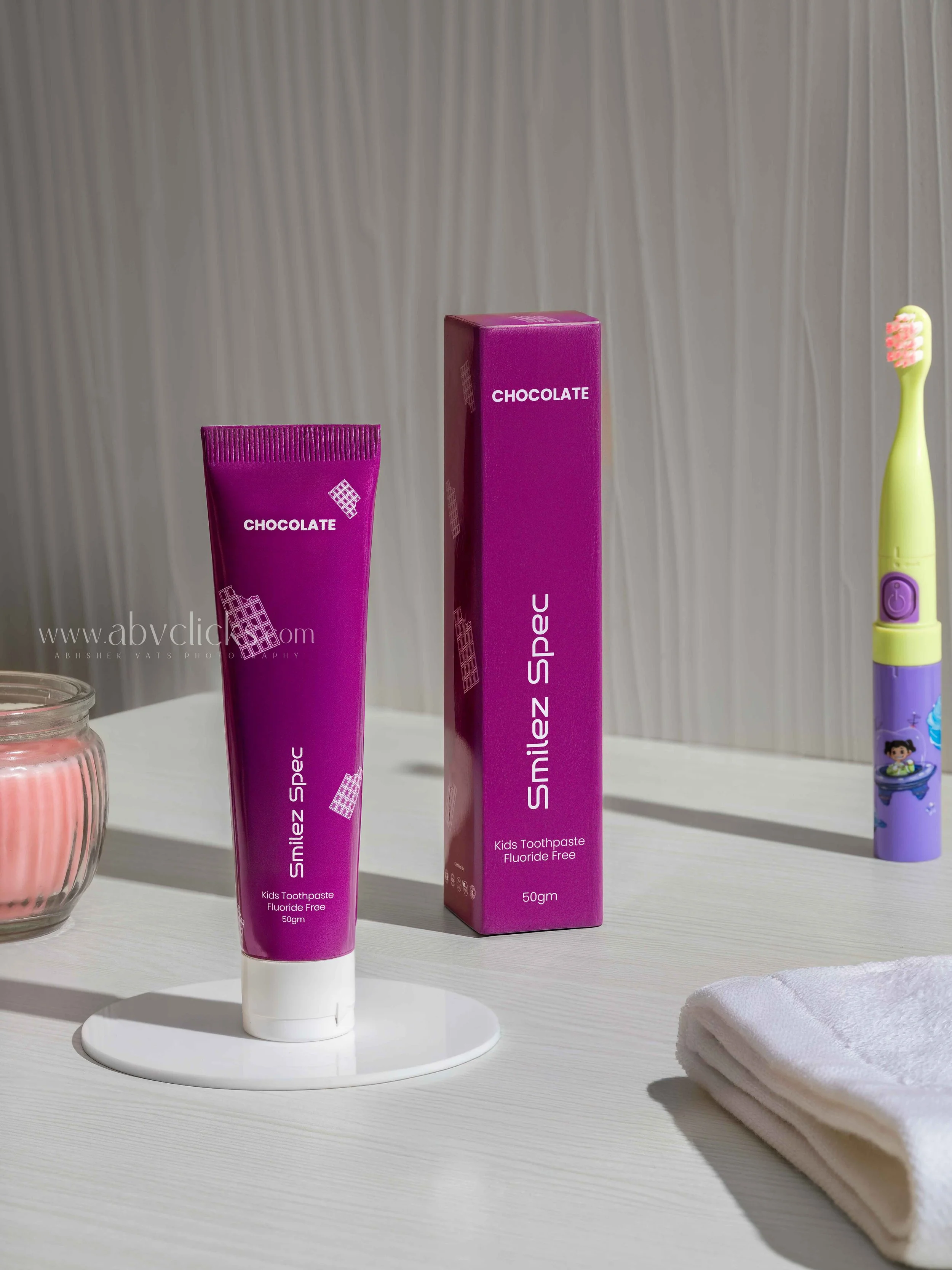 Pink kids toothpaste tube labeled "Smilez Spce" placed on a white stand, with a matching boxed toothpaste behind it, on a white countertop. To the right, there is a colorful children's electric toothbrush and a neatly folded white towel. A small glas