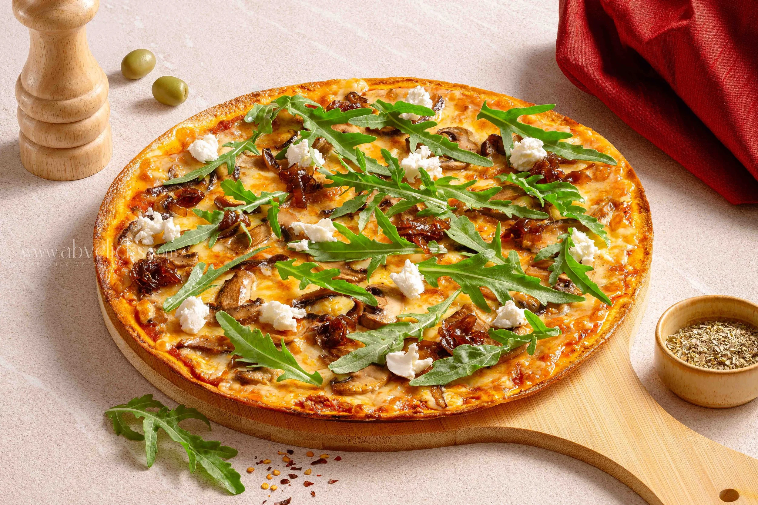 Pizza topped with arugula, sun-dried tomatoes, cheese, and mushrooms on a wooden serving board. There is a red napkin, a wooden pepper grinder, and bowls of herbs and seasoning nearby.