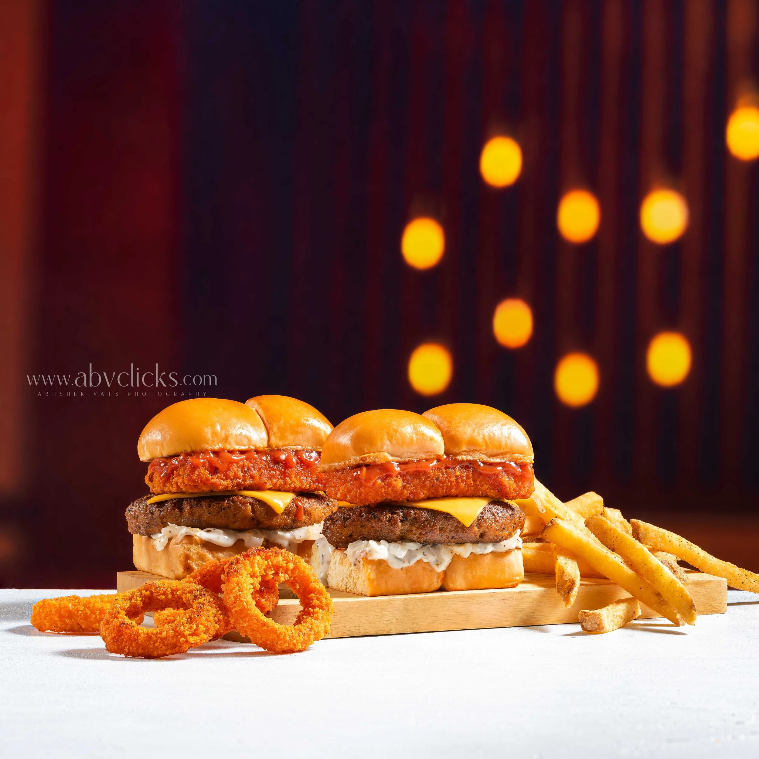 A wooden serving board with three stacked cheeseburgers and fried chicken strips with curly fries on the side. The background is dark with blurred warm-colored lights.