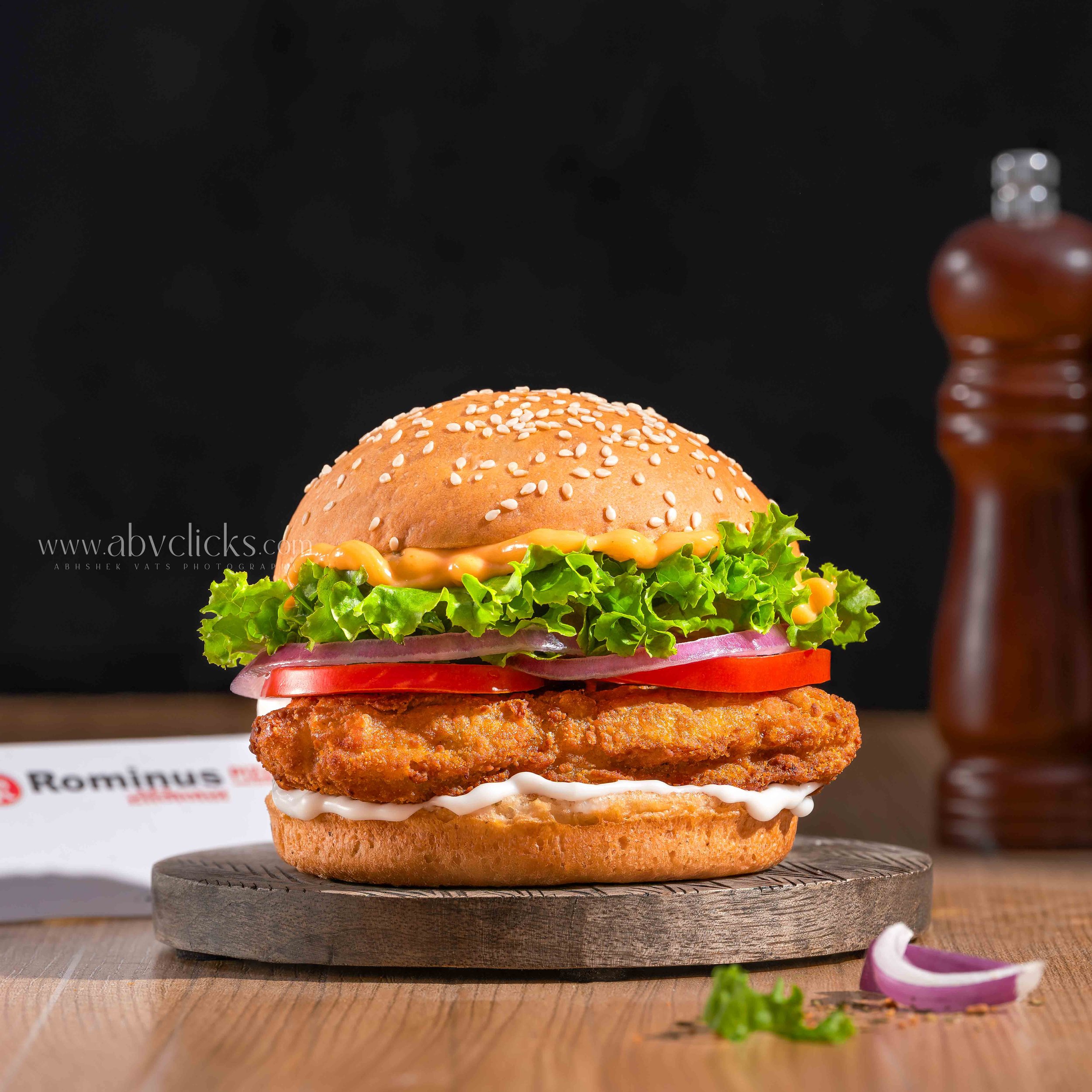 A burger with lettuce, tomato, cheese, fried chicken patty, onion, and mayonnaise inside a sesame seed bun, placed on a wooden board.