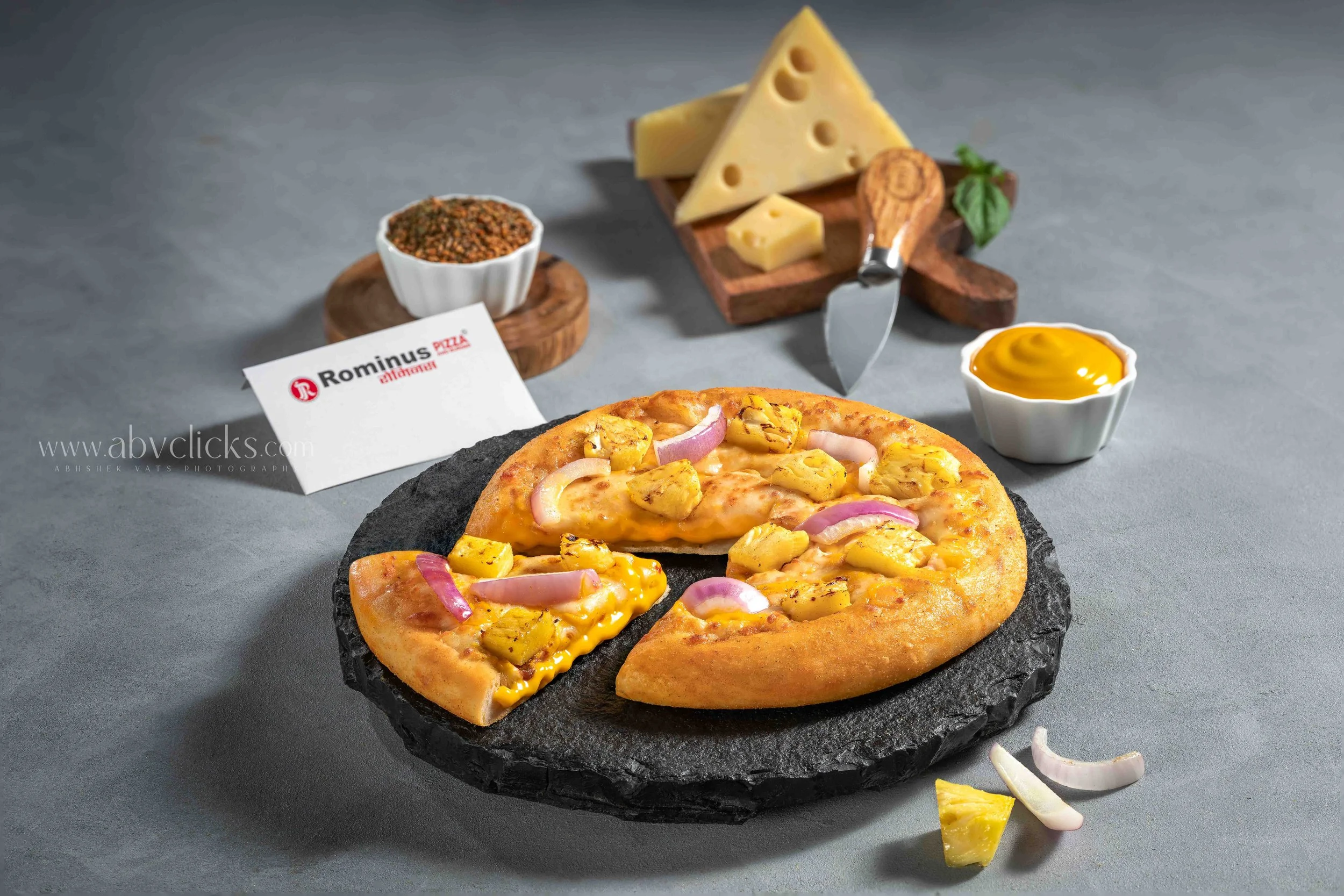 Close-up of a pizza with pineapple chunks and red onion slices on a black slate serving board. In the background, there are slices of yellow cheese, a small bowl of oregano, a small bowl of honey, and a block of cheese with a cheese cutter on top. Th