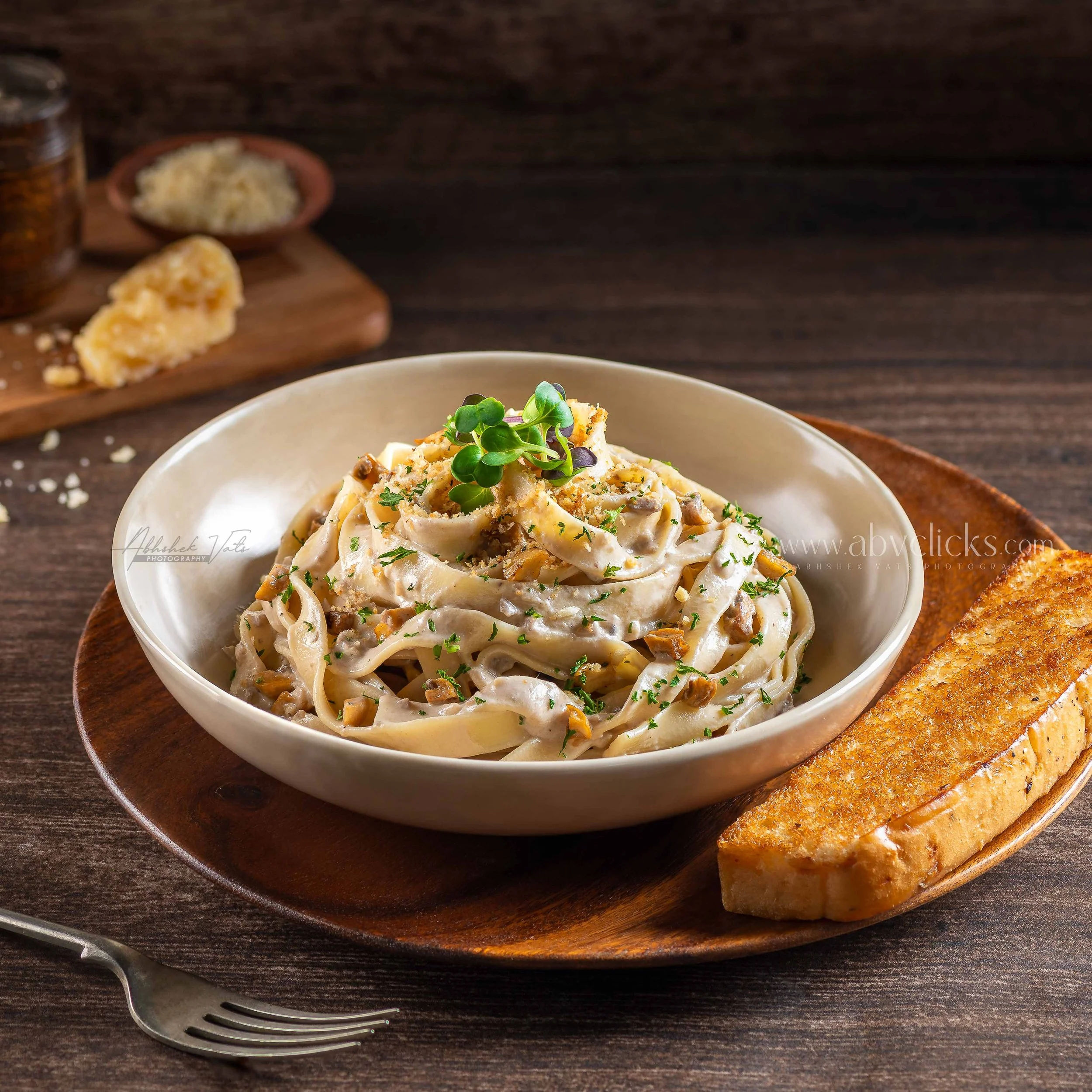Creamy pasta with mushrooms topped with microgreens on a white plate, placed on a wooden tray, with garlic bread and grated cheese in the background on a dark wood table.