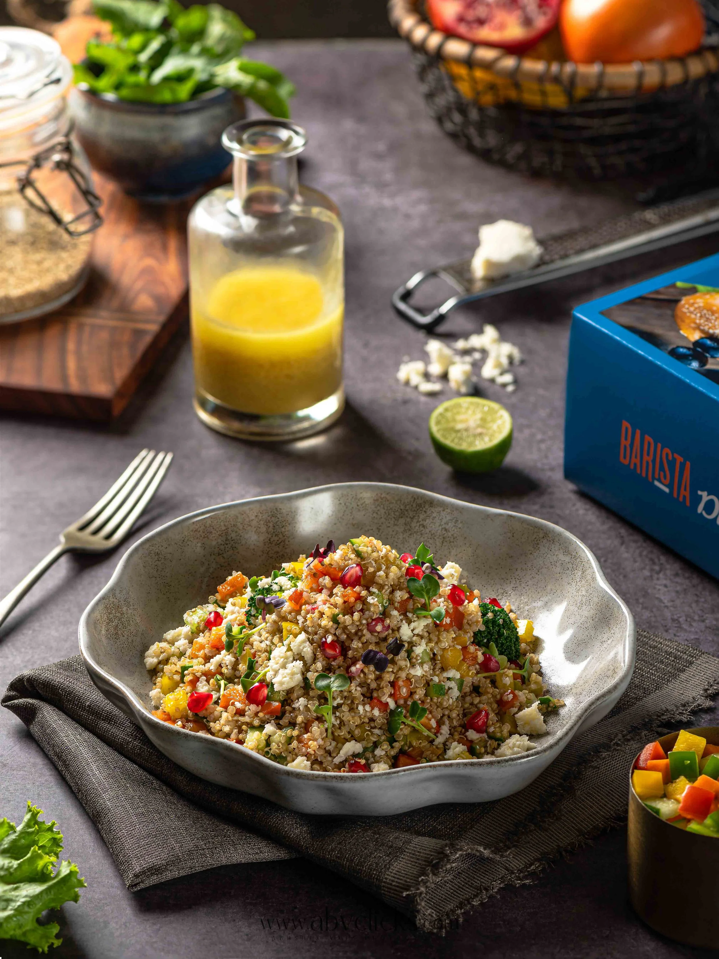A bowl of quinoa salad with chopped vegetables and garnished with microgreens, set on a dark table with a fork, lime half, and various other dishes and ingredients in the background.