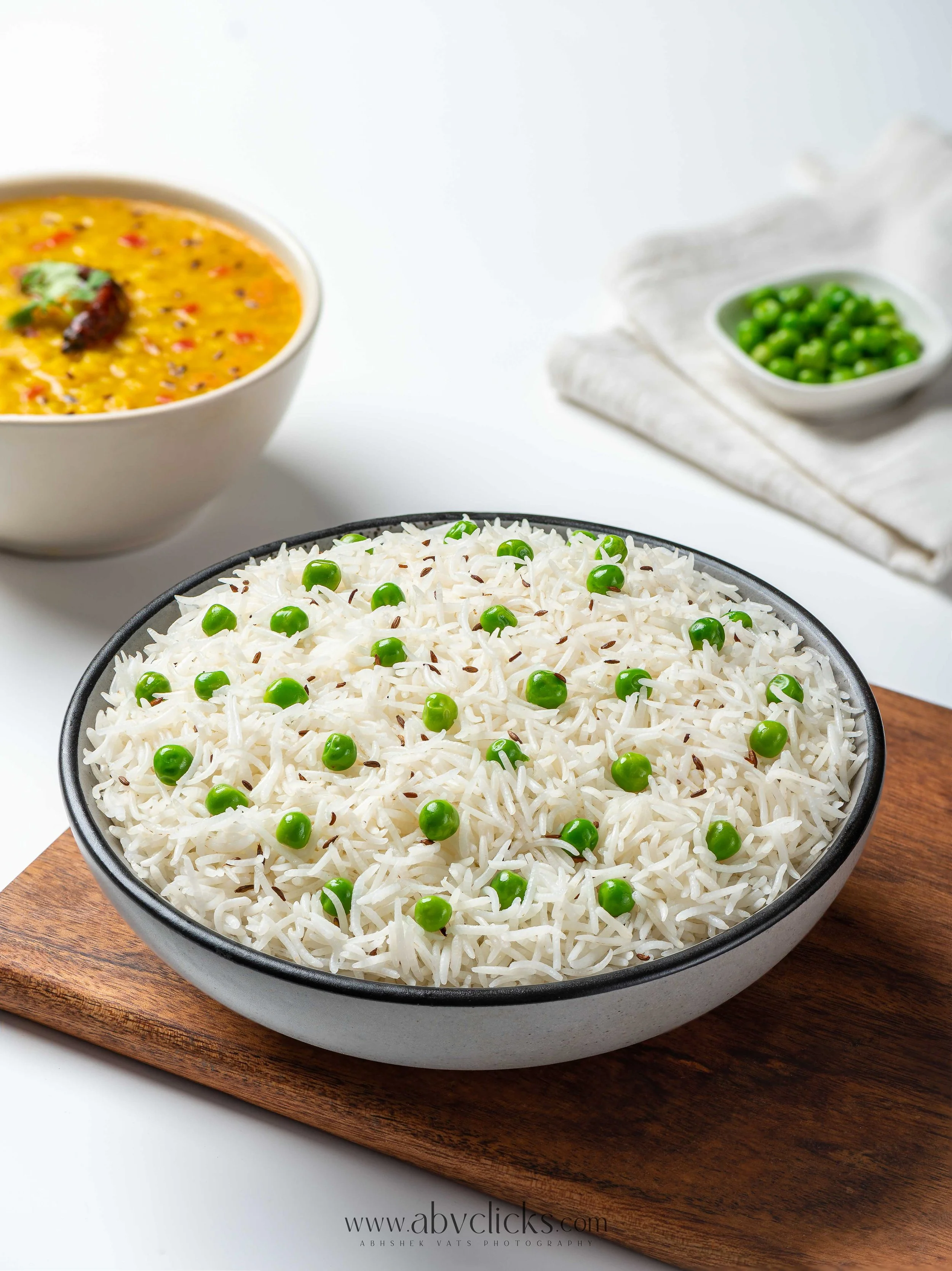 A black bowl of cooked rice with green peas on top, placed on a wooden board. In the background, a bowl of yellow lentil soup with red chili flakes and herbs, and a small white dish of green peas are visible.
