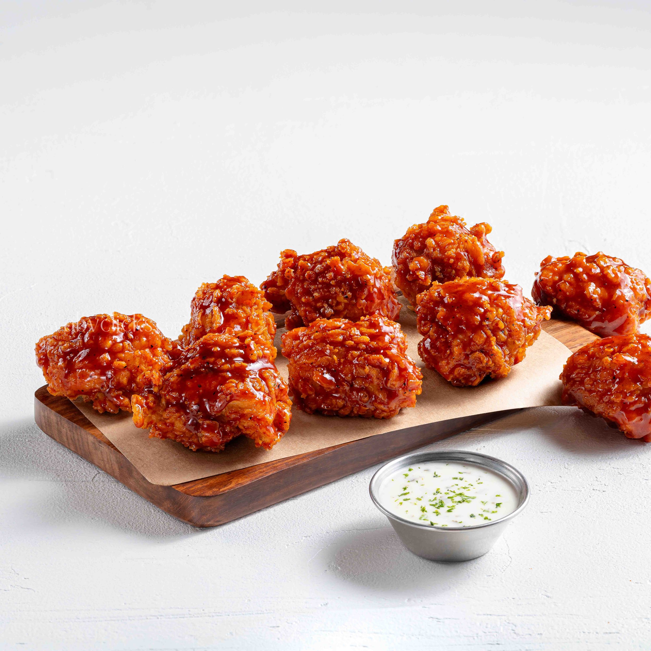 A wooden serving board with several pieces of glazed fried chicken on a white surface, accompanied by a small bowl of creamy white dipping sauce garnished with chopped herbs.
