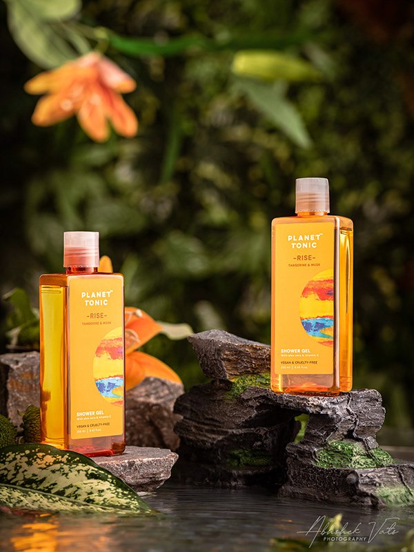Two bottles of Planet Tonic Rise shower gel on stacked rocks outdoors, with greenery and orange flowers in the background.