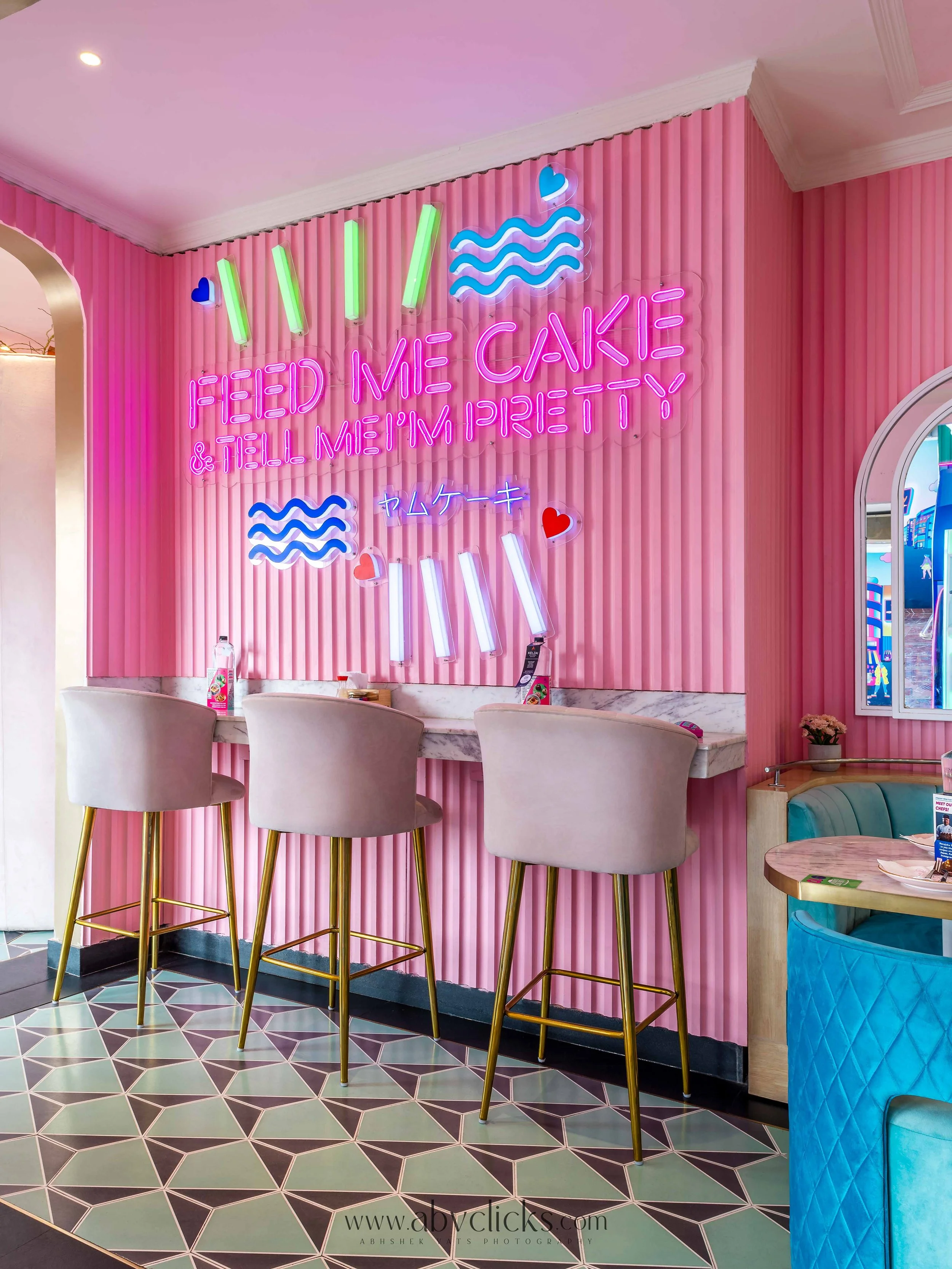 Interior of a pink-themed cafe with a pink striped wall, neon signs, and white bar stools with gold legs situated at a marble counter. The decor includes neon lights with phrases and symbols in bright colors, and a patterned tile floor.