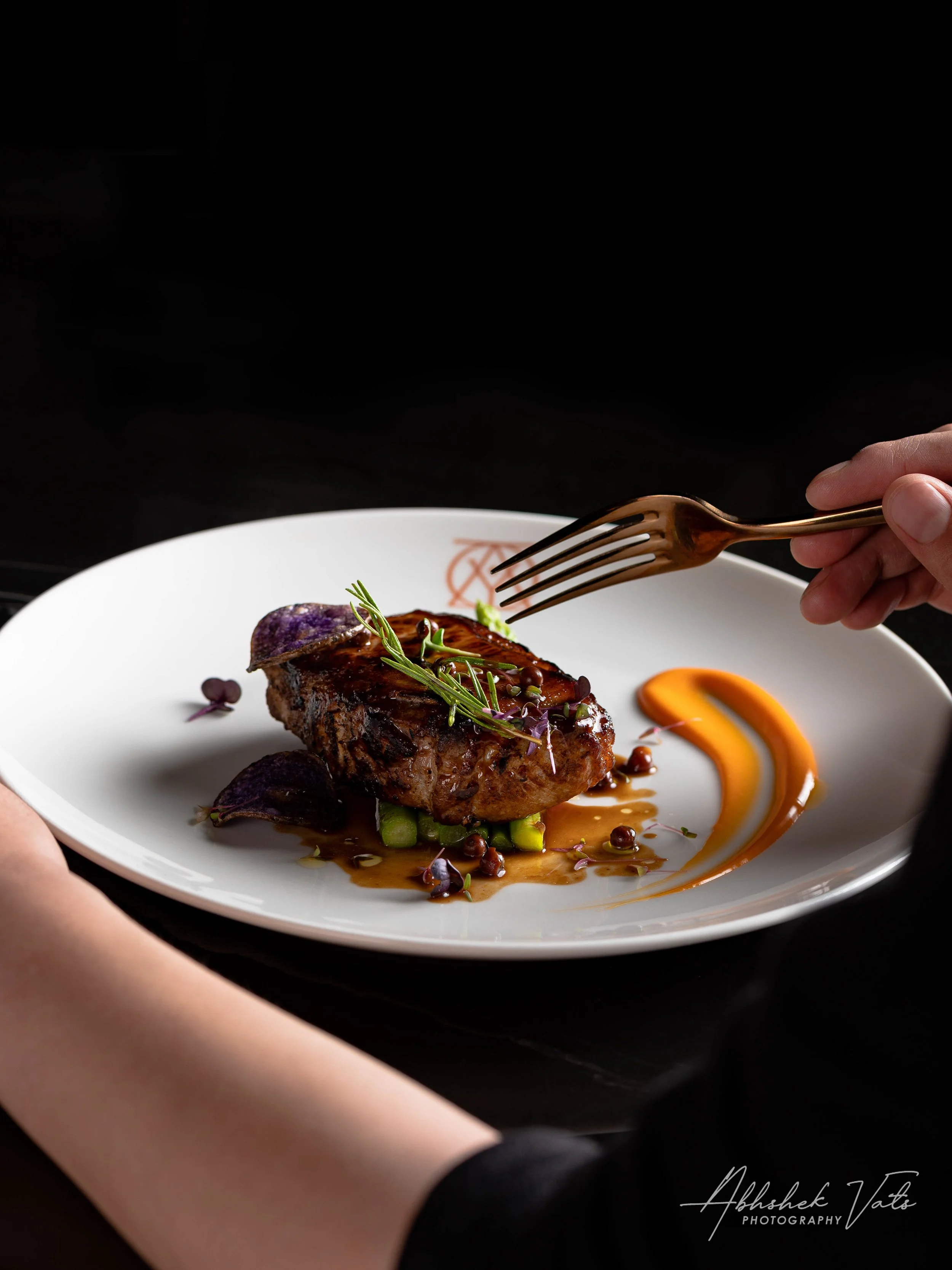 A cooked piece of meat garnished with herbs on a white plate, decorated with purple vegetables and orange sauce swirl, with someone's hand holding a fork above.