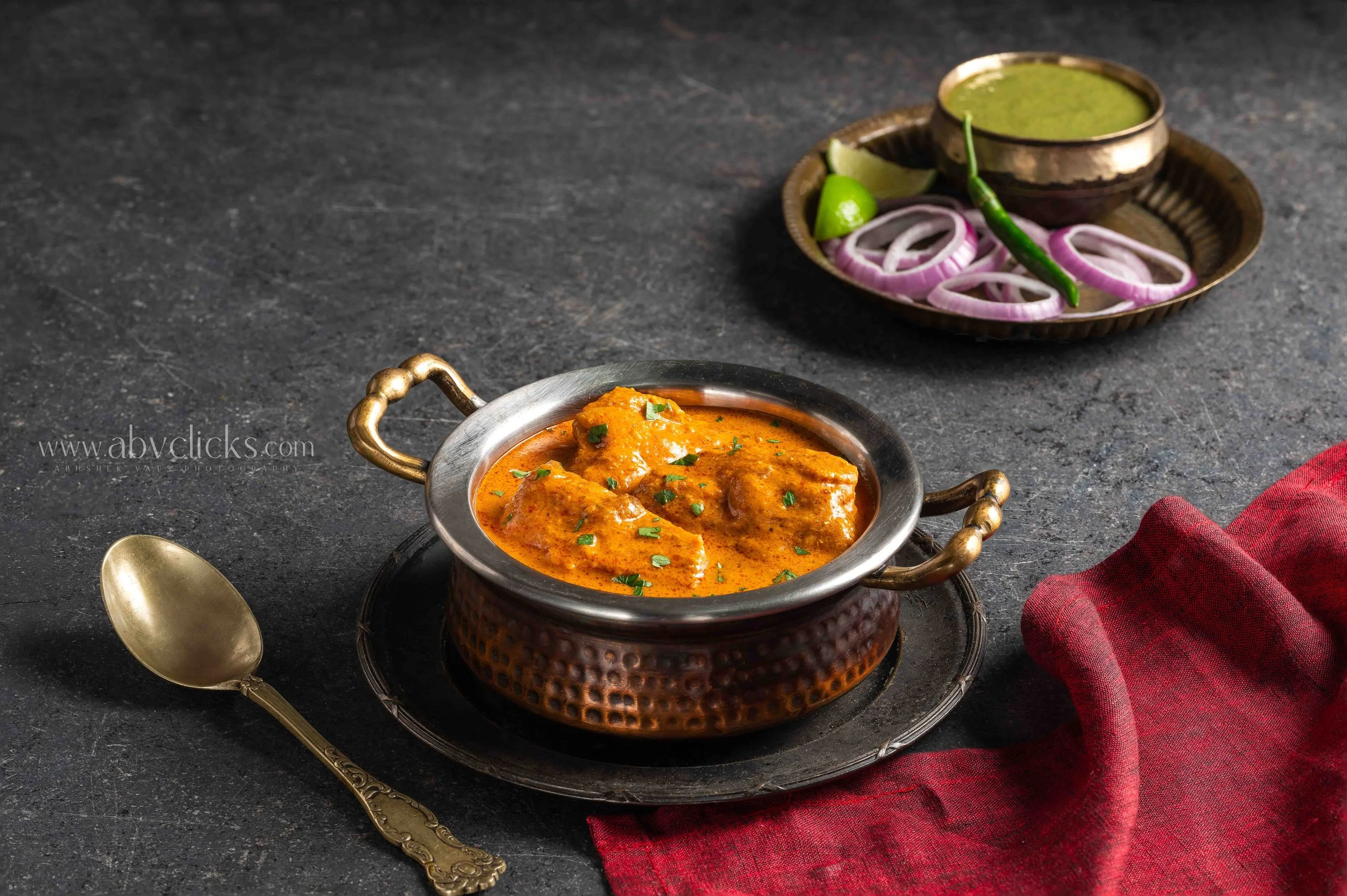 A plate of Indian chicken curry garnished with herbs, a bowl of green chili sauce, lime wedges, sliced onions, a gold spoon, and a red cloth napkin on a dark surface.