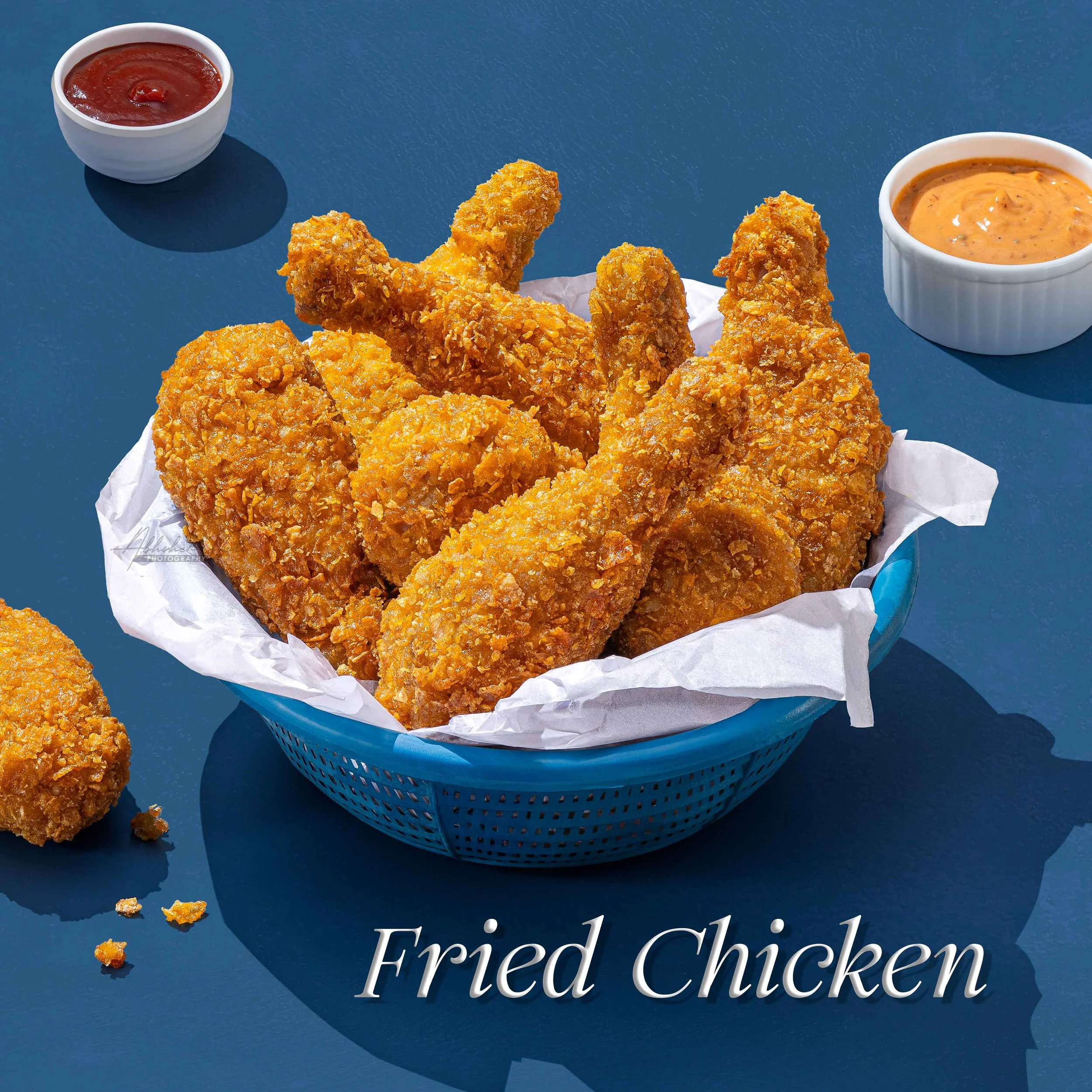 Basket of fried chicken pieces with two side sauces, ketchup on the left and spicy mayonnaise on the right, on a blue surface with scattered fried chicken crumbs and partially visible text reading 'Fried Chicken'.