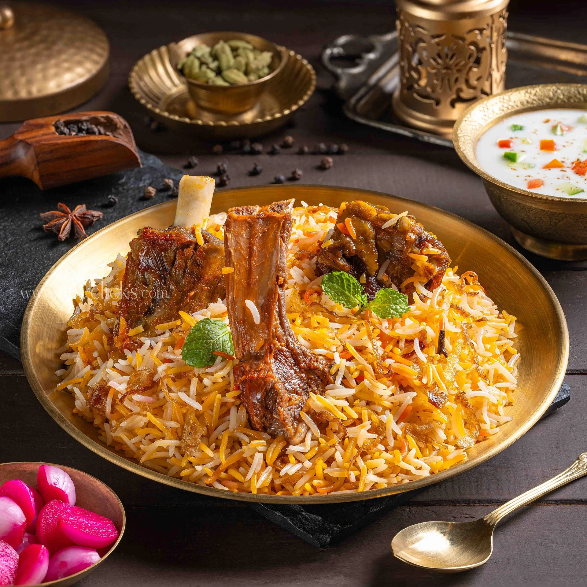 A close-up of a plate of Indian chicken biryani garnished with cilantro leaves, with a bowl of vegetable raita and a smaller bowl of green cardamom pods in the background, and pink and white pickled radishes in a separate bowl in the foreground, on a