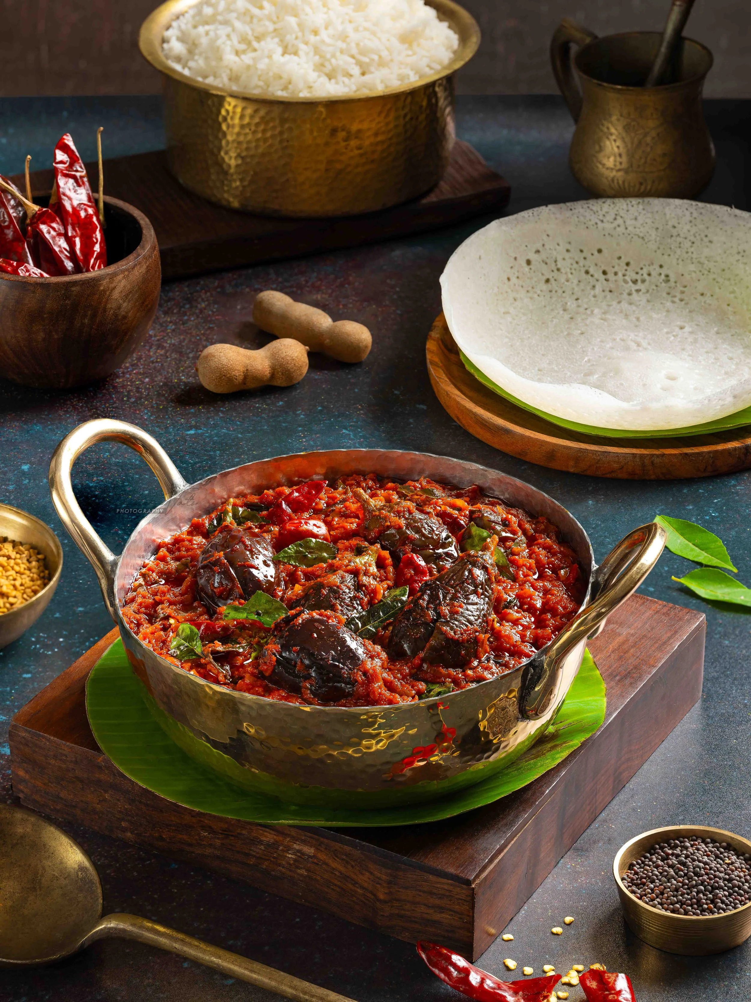 A bowl of spicy eggplant curry garnished with curry leaves on a banana leaf, surrounded by spices including dried red chilies, peppercorns, and mustard seeds, with white rice in a brass bowl in the background.