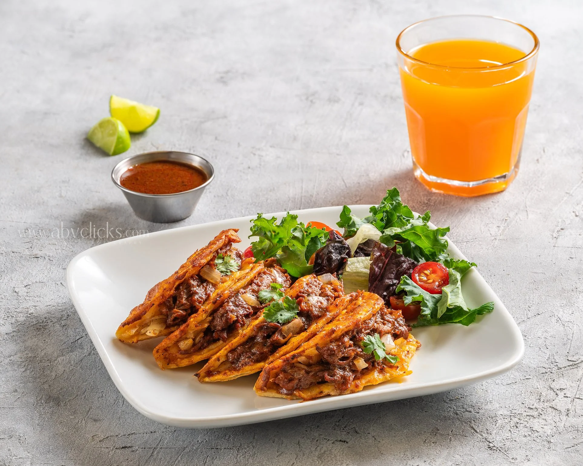 Plate of Mexican food topped with shredded meat, cheese, and fresh salad, with a side of hot sauce, lime wedges, and a glass of orange juice on a gray surface.