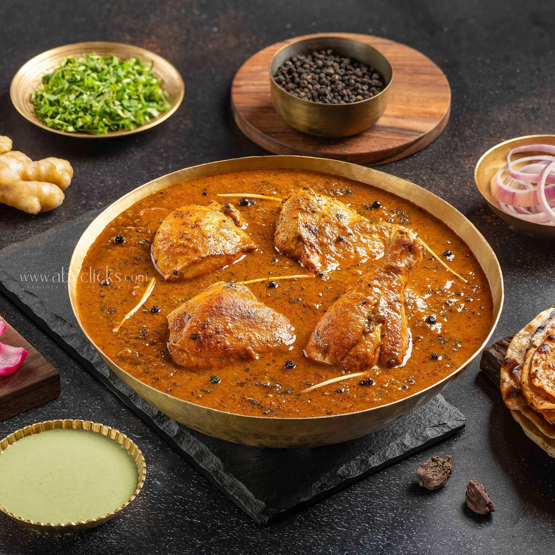 A serving of chicken cooked in a rich, spicy Indian curry in a golden bowl, garnished with black peppercorns. Surrounding the curry are small bowls with chopped green herbs, black peppercorns in a wooden container, sliced red onions, and a green sauc