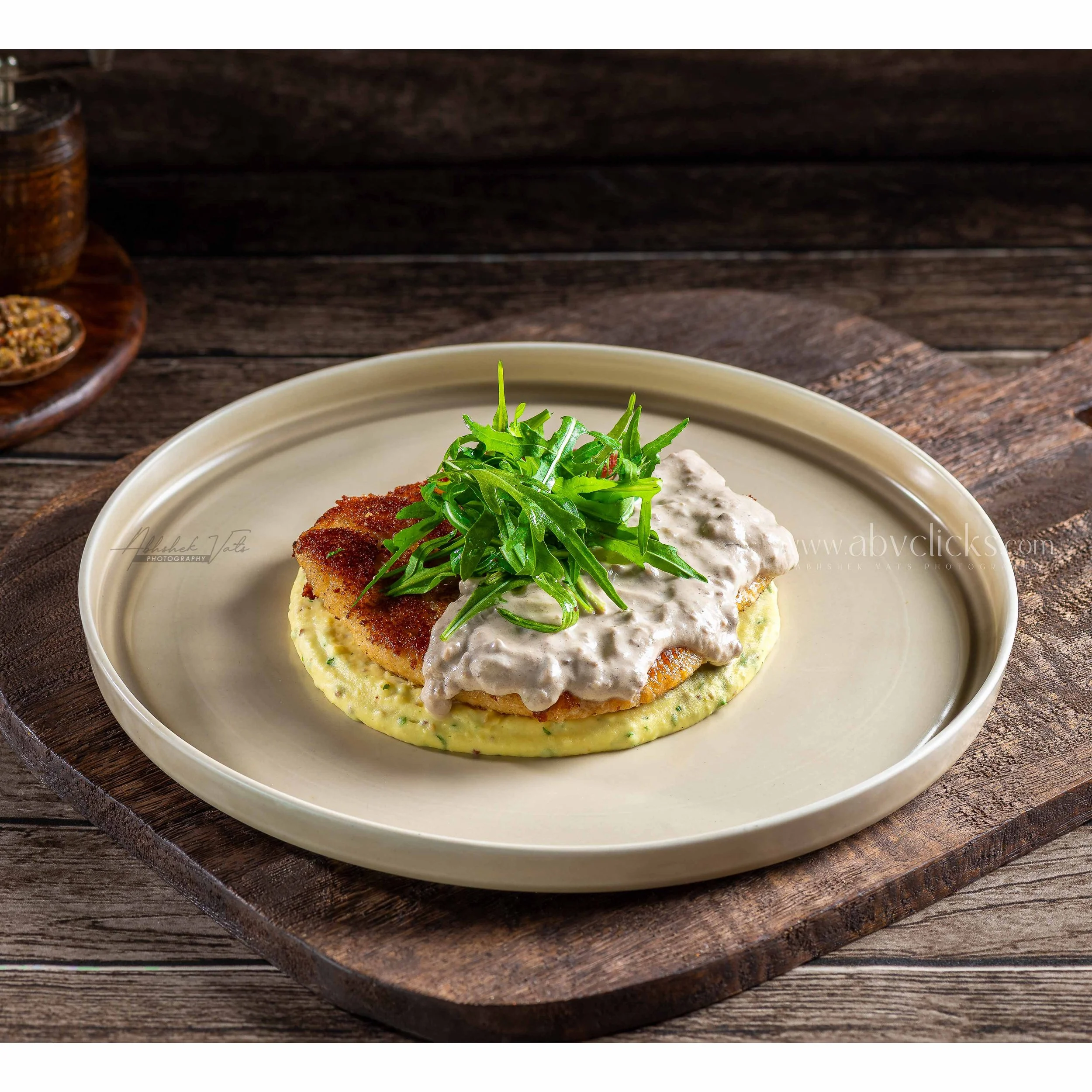 A plate with a breaded meat cutlet topped with fresh green arugula, creamy white sauce, and mashed potatoes, set on a wooden surface.