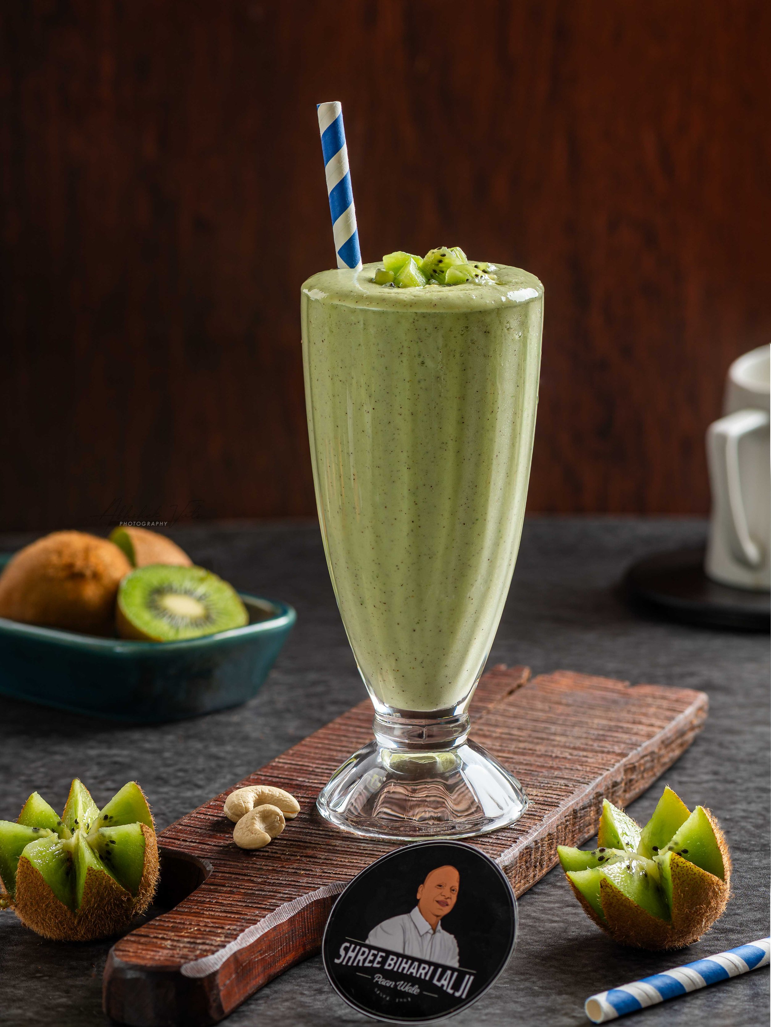 A tall glass of green smoothie garnished with chopped kiwi and chia seeds, with a blue striped straw. Surrounding the glass are halved kiwis, cashews, and a small black circular sign with a man’s portrait and the text 'SHREE BIHARILALJI Peace Upload'