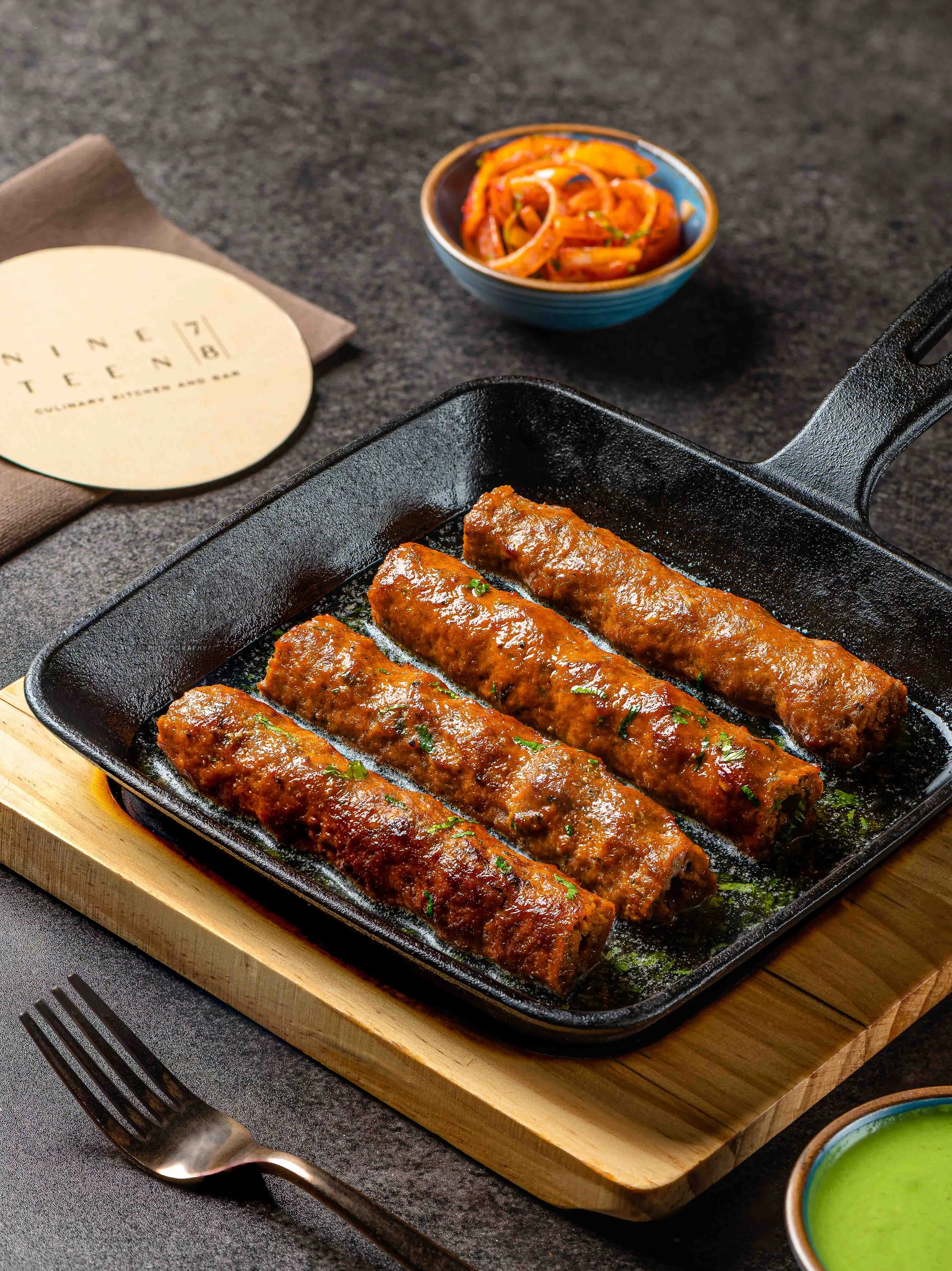 Three glazed sausage links in a cast iron skillet on a wooden board. A bowl of spicy sliced onions and peppers, a menu, and small bowl of green sauce are visible nearby.
