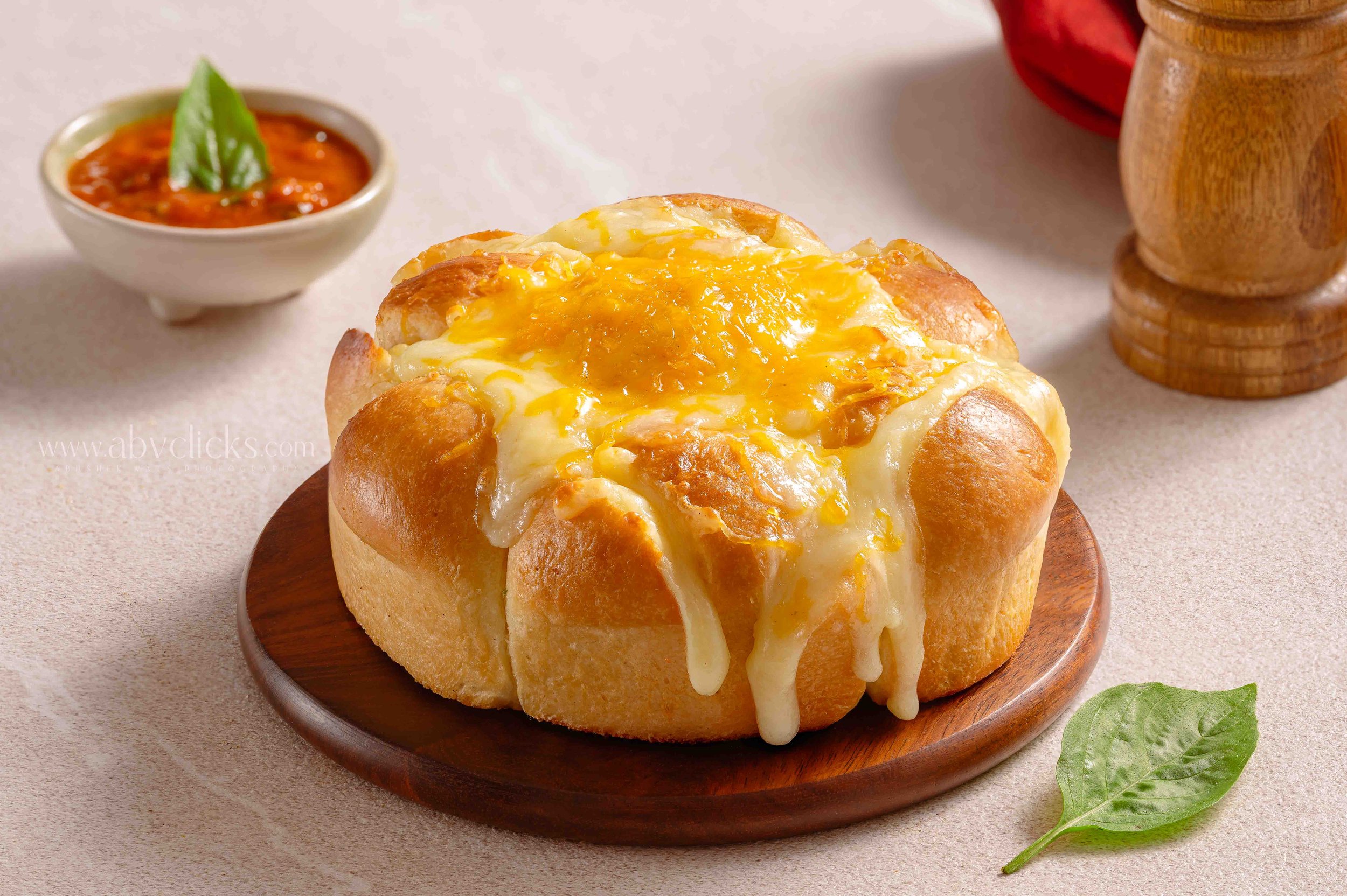 A bread bowl filled with melted cheese and egg, with cheese dripping over the sides, placed on a wooden plate. In the background, there is a small bowl of red sauce with a green garnish and a wooden pepper grinder.