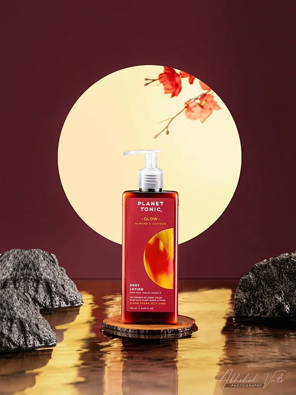 A bottle of Planet Tonic glow body lotion with almond and saffron scent, placed on a small wooden platform amidst rocks, with a large illuminated moon and red flowers in the background.