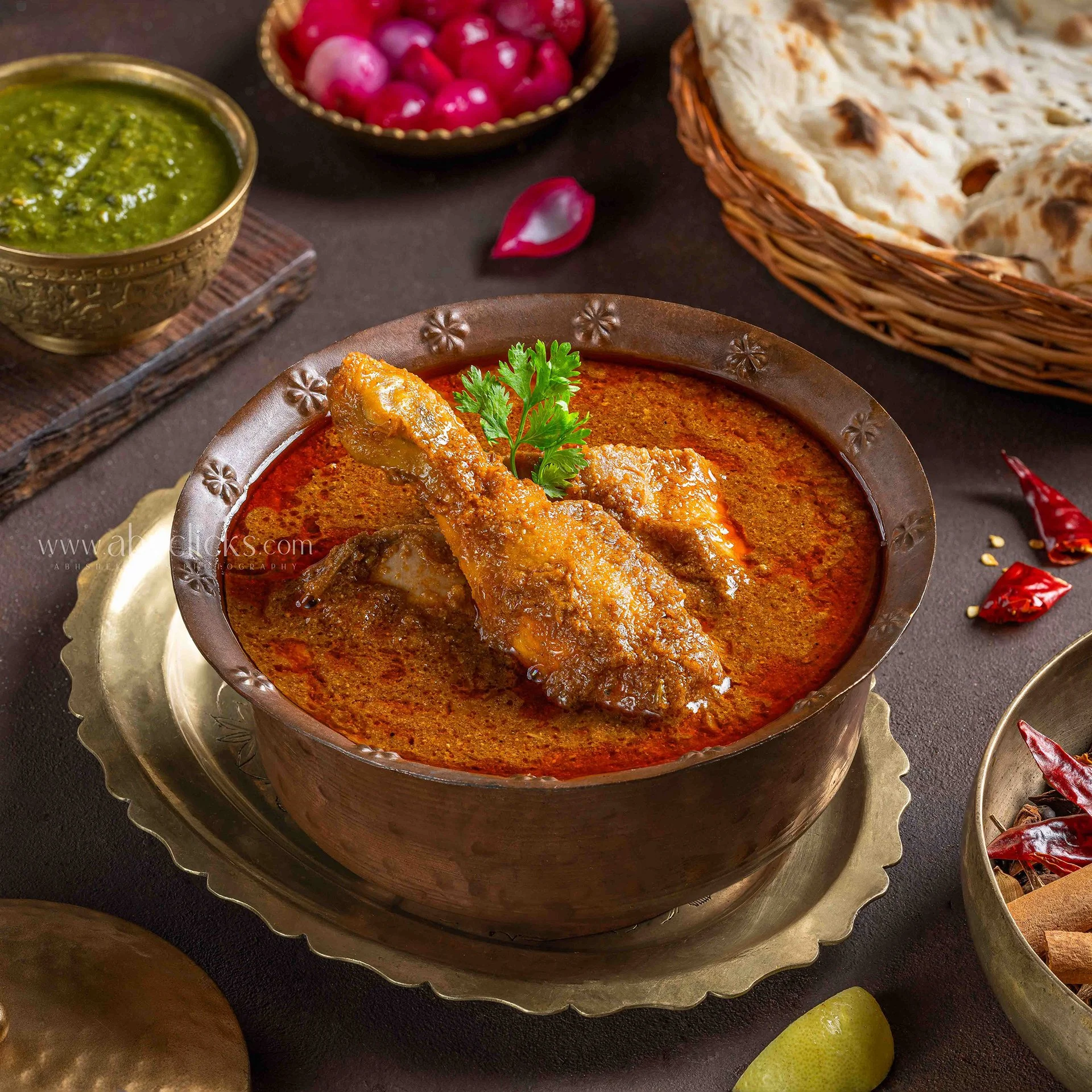 Indian cuisine spread with a bowl of chicken curry garnished with cilantro, green chutney in a decorative bowl, fresh radishes in a bowl, papadums in a wicker basket, chili peppers, a lemon, and a salt shaker on a dark surface.