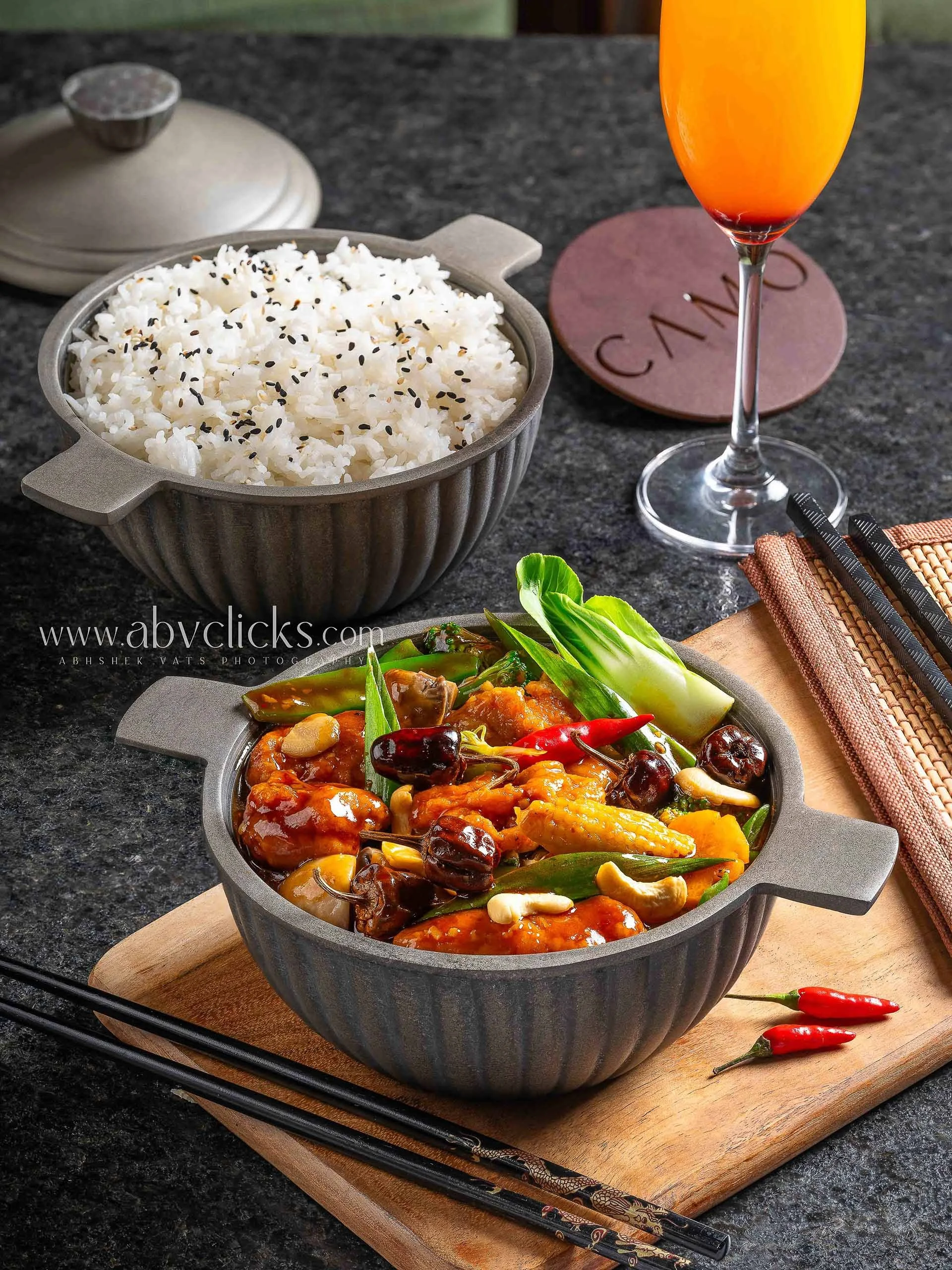 A bowl of white rice sprinkled with black sesame seeds, a colorful vegetable dish with sauce in a gray cast iron skillet, a glass of orange-colored drink, chopsticks, a small container of spices, and a red chili pepper on a wooden cutting board.